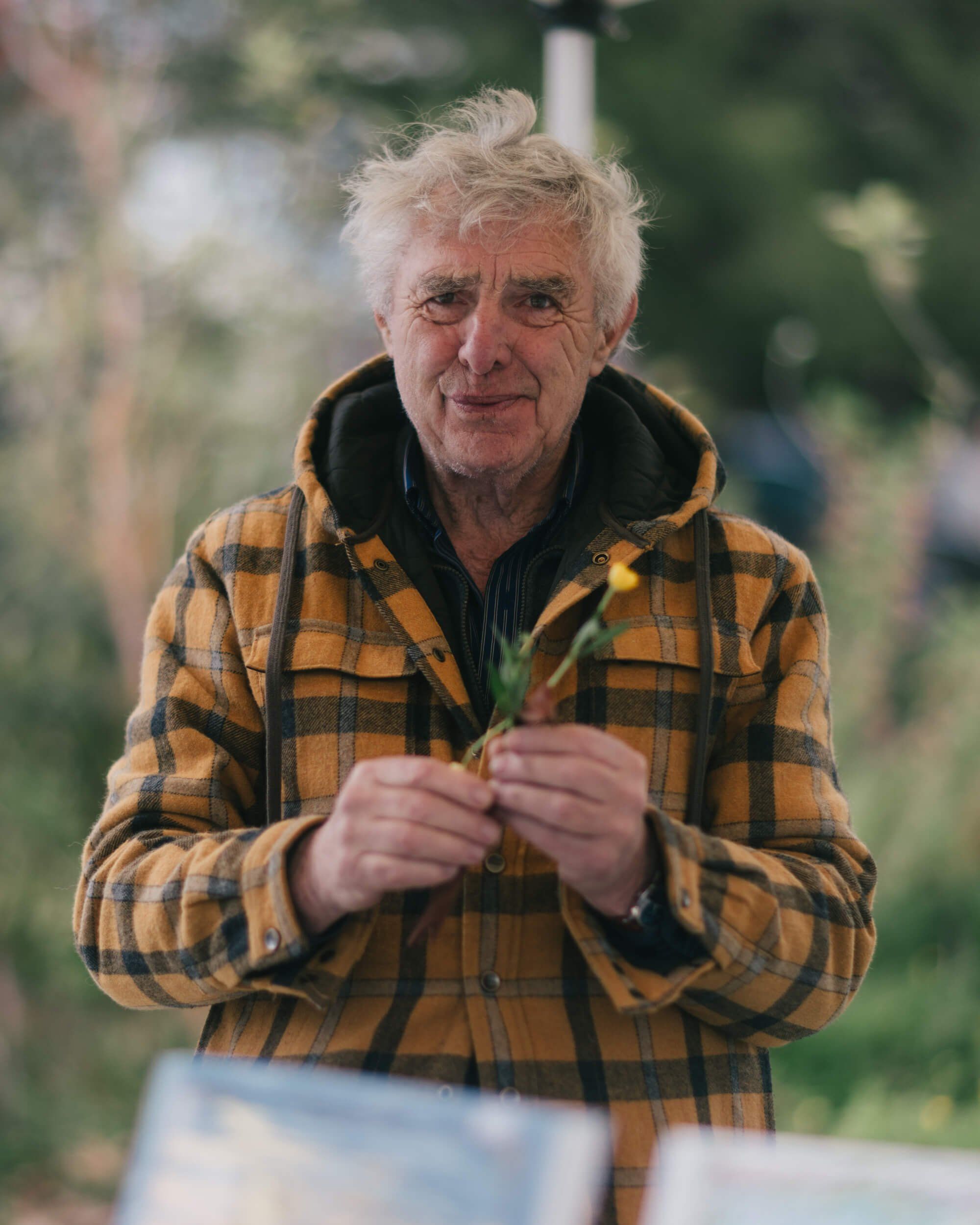Older man with white hair in yellow plaid shirt holds seedlings at outdoor market, soft natural light, blurred green foliage background
