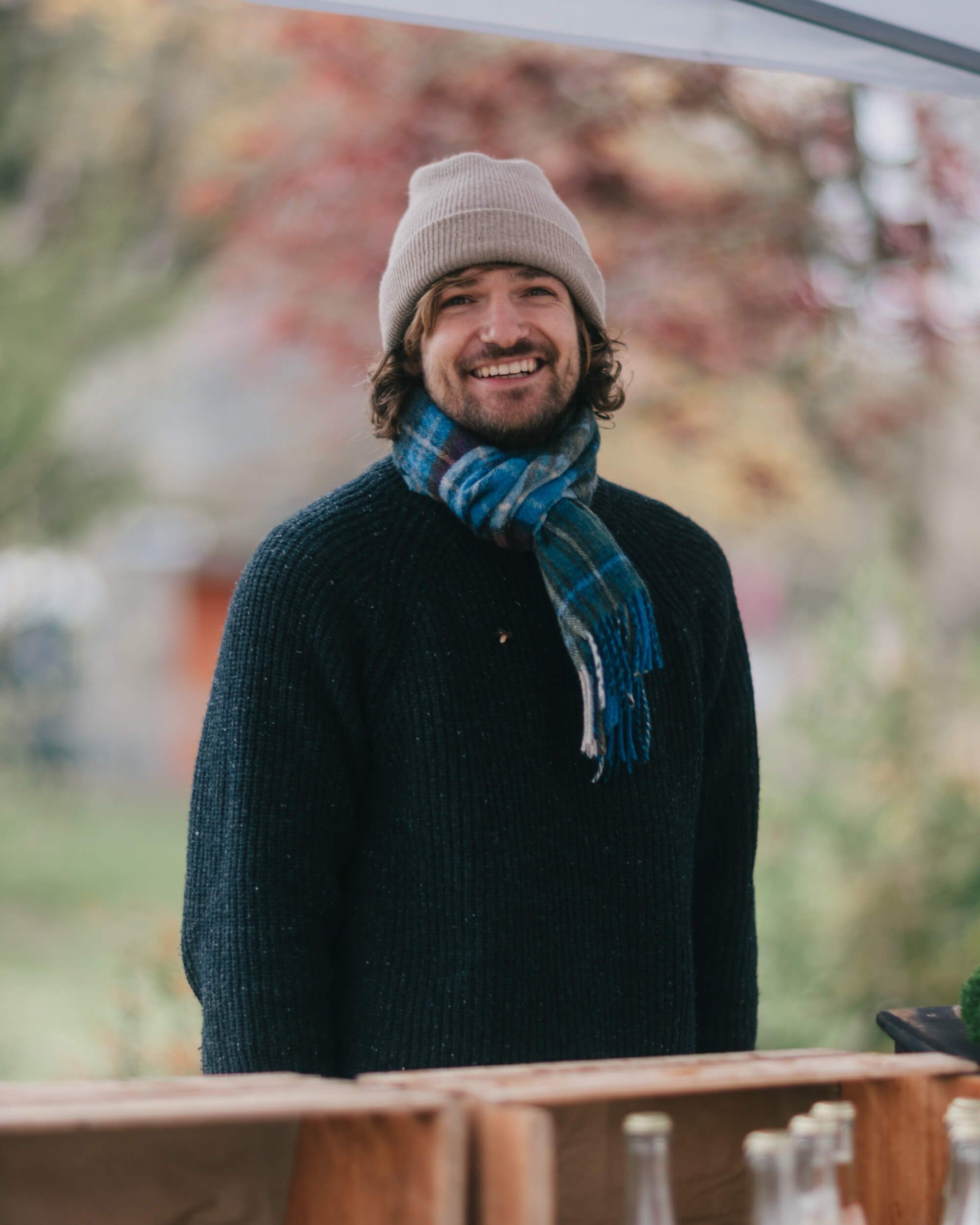 Smiling man in cream beanie and blue plaid scarf leans against wooden railing with autumn foliage softly blurred behind him