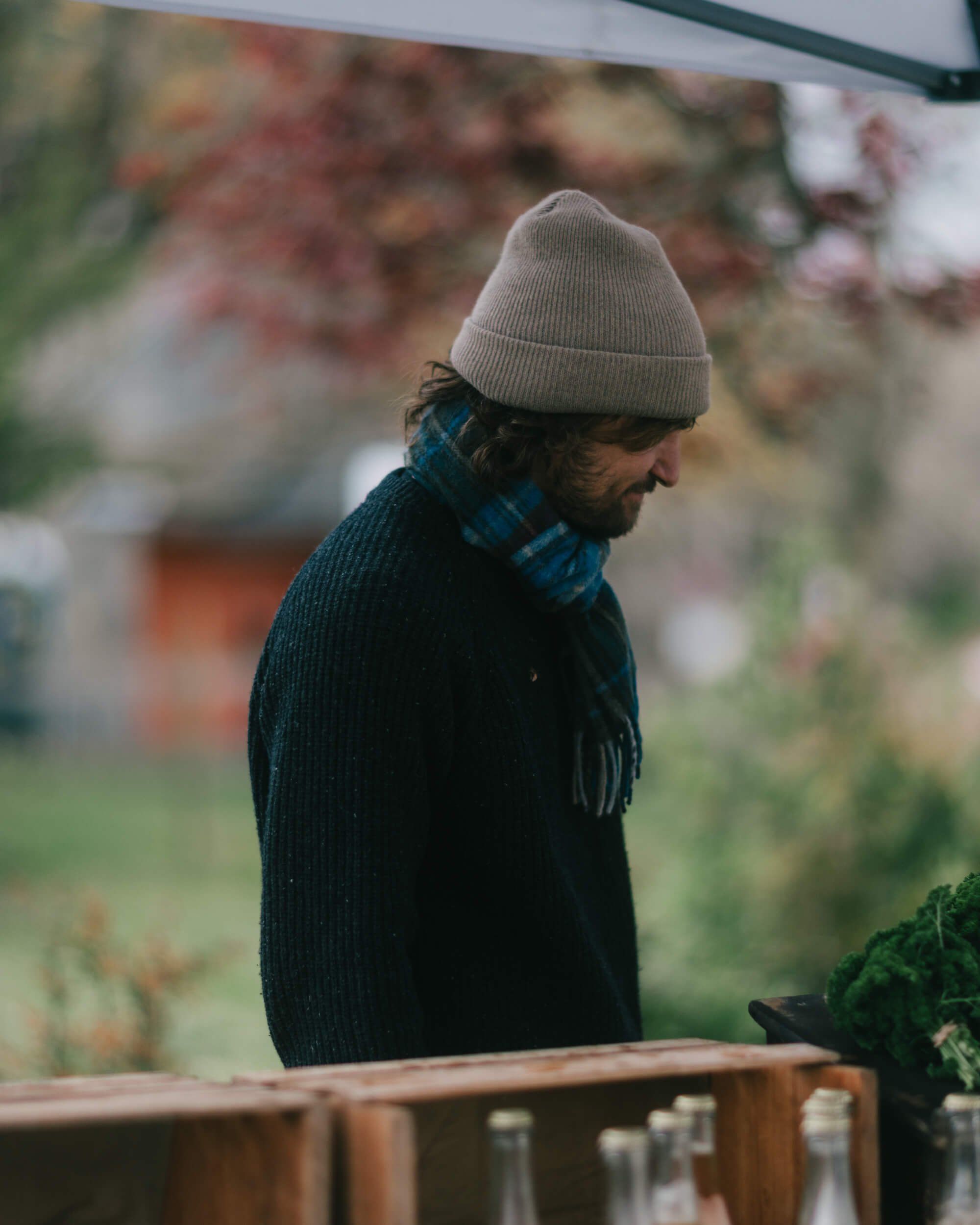 Bearded man in cream beanie and dark sweater gazes downward at Salt Spring Island Saturday Market, soft autumn light filtering through blurred foliage behind