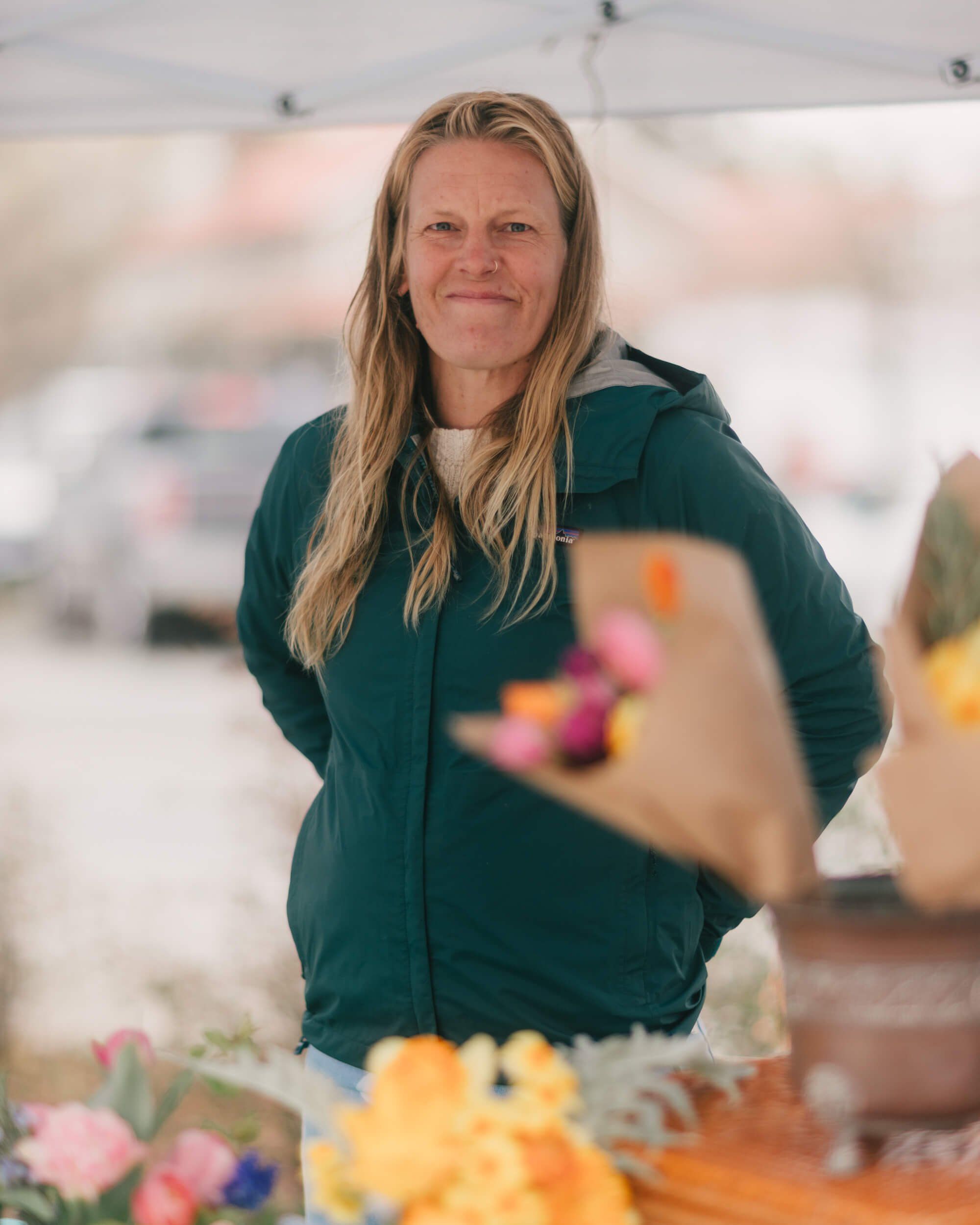 Woman in dark teal hoodie smiles at Saturday Market among soft-focus flowers and vendor tents