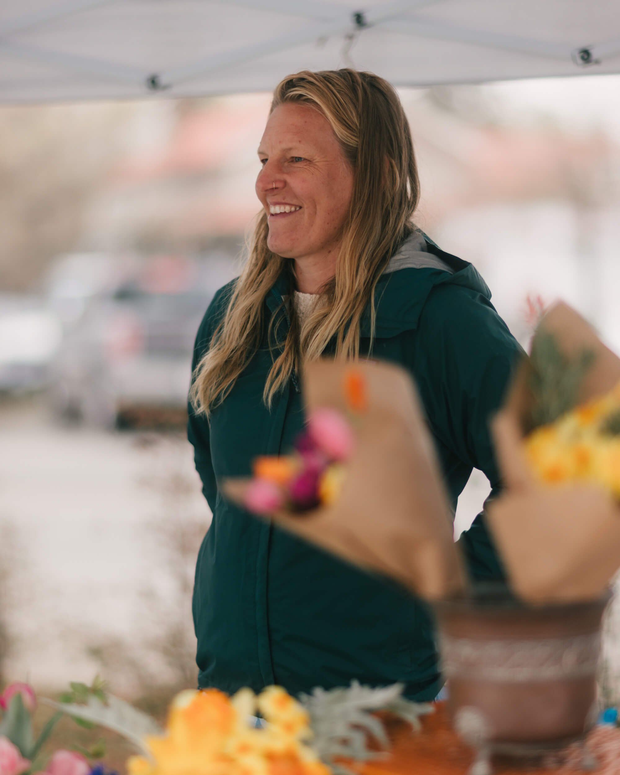 Woman in dark teal jacket smiles among colorful potted flowers at outdoor market under tent