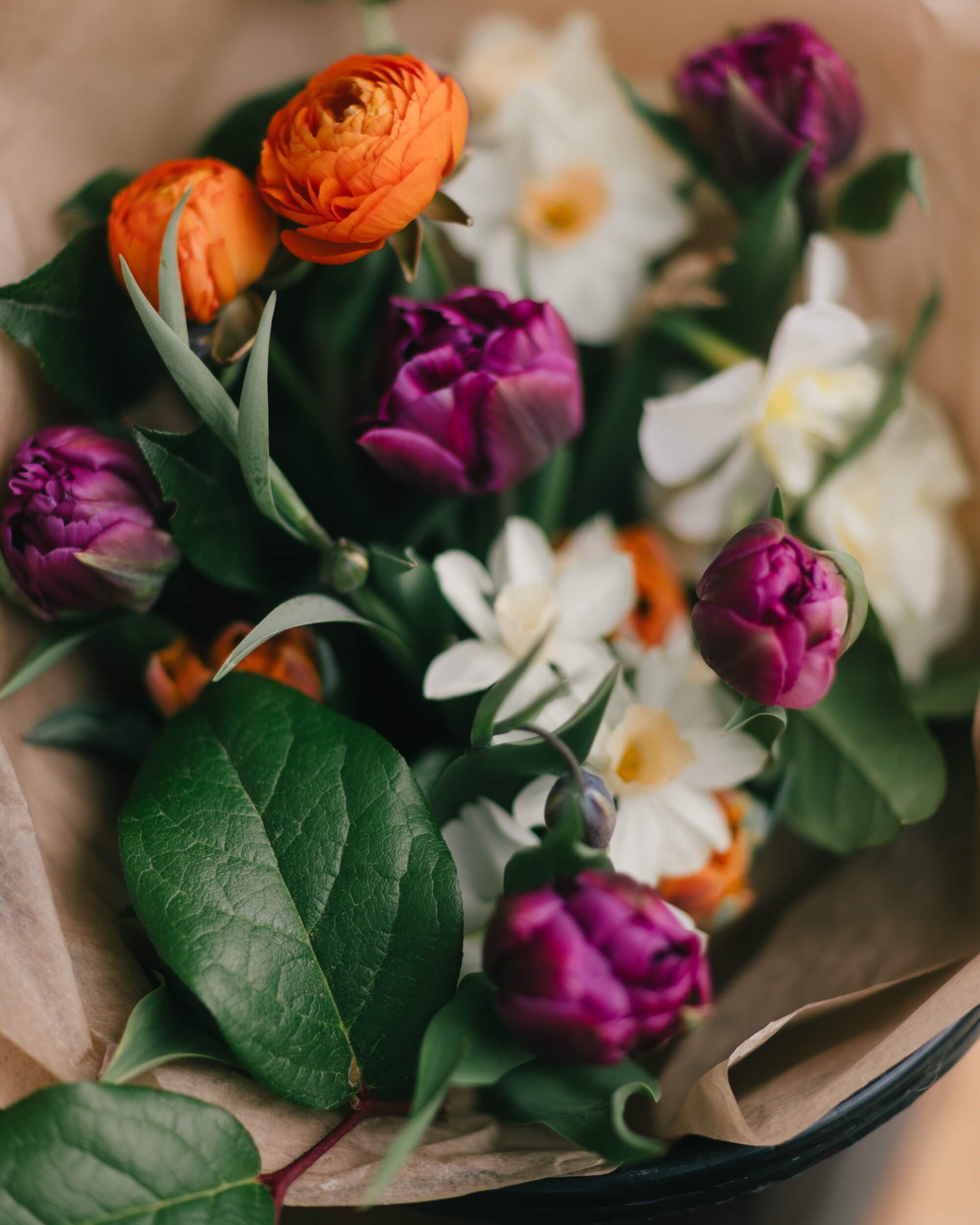 Vibrant bouquet of orange ranunculus, deep purple tulips, and white daisies wrapped in kraft paper, held against a warm neutral background