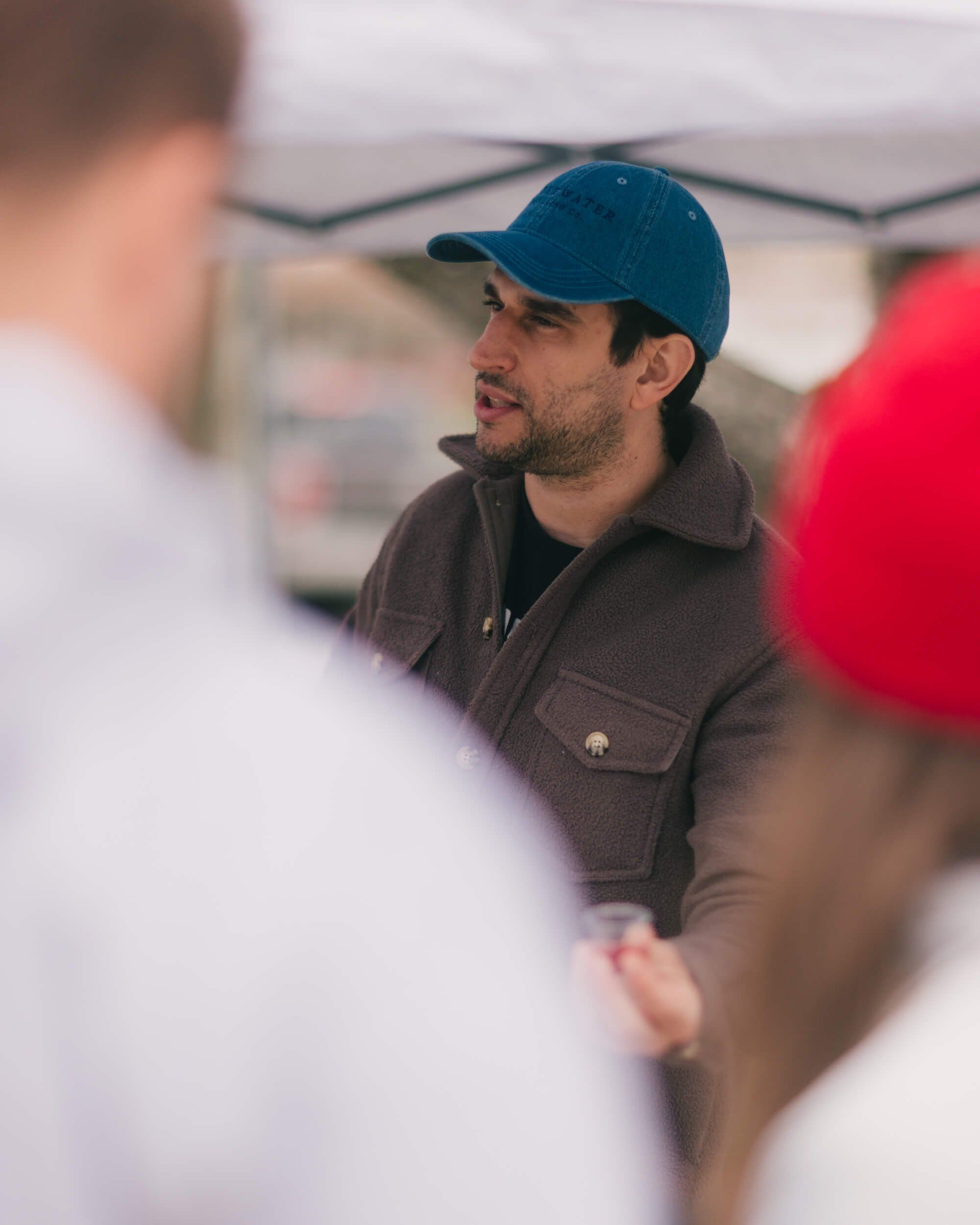 Man in blue cap and charcoal jacket smiles while conversing at an outdoor market under a tent canopy