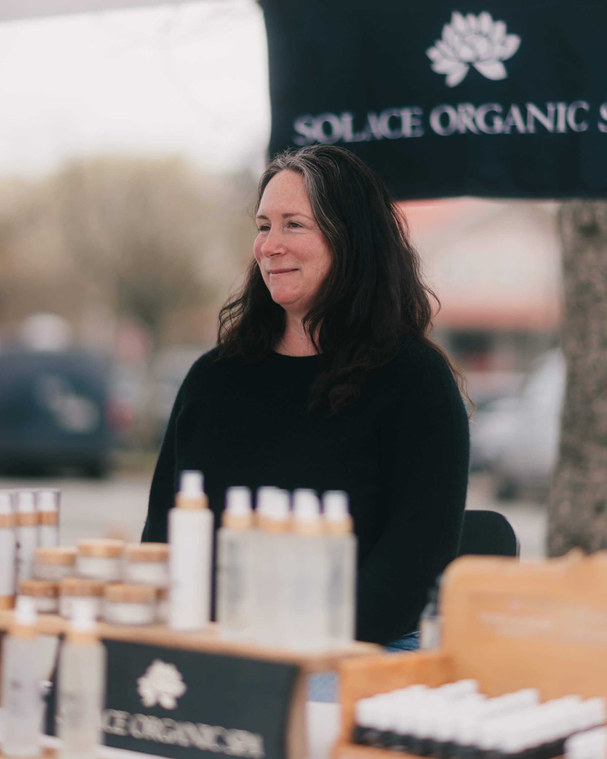 Woman with long dark hair smiling at an outdoor market booth displaying organic skincare products in white bottles