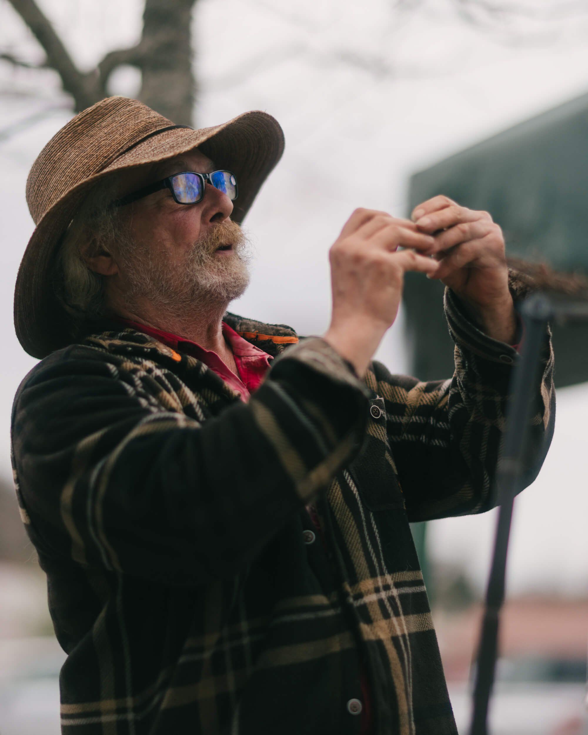 Man in plaid shirt and cowboy hat playing harmonica at outdoor market, blue-tinted sunglasses, overcast sky