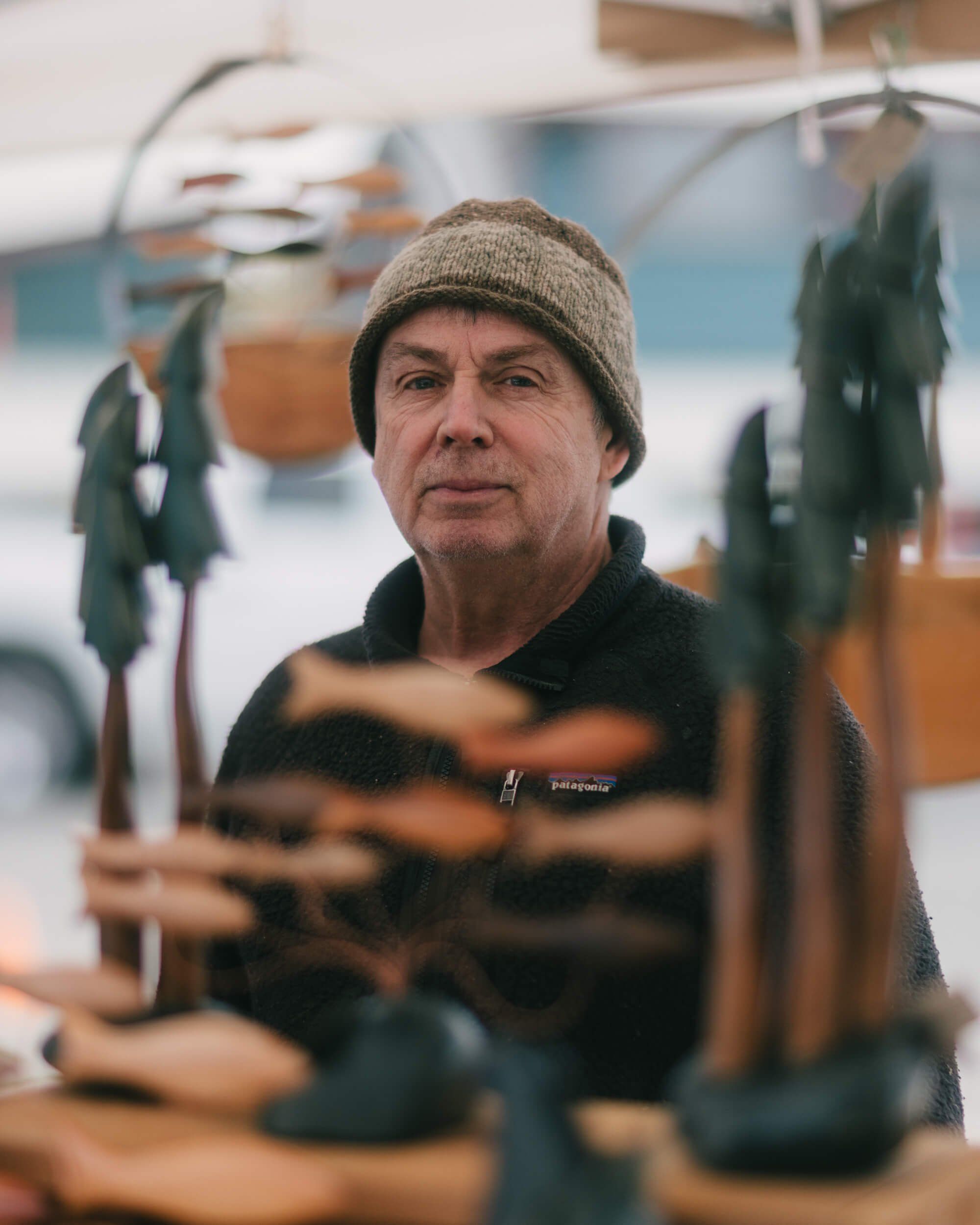 Focused artisan among handcrafted wooden pieces at Saturday Market, wearing a knit beanie and dark jacket