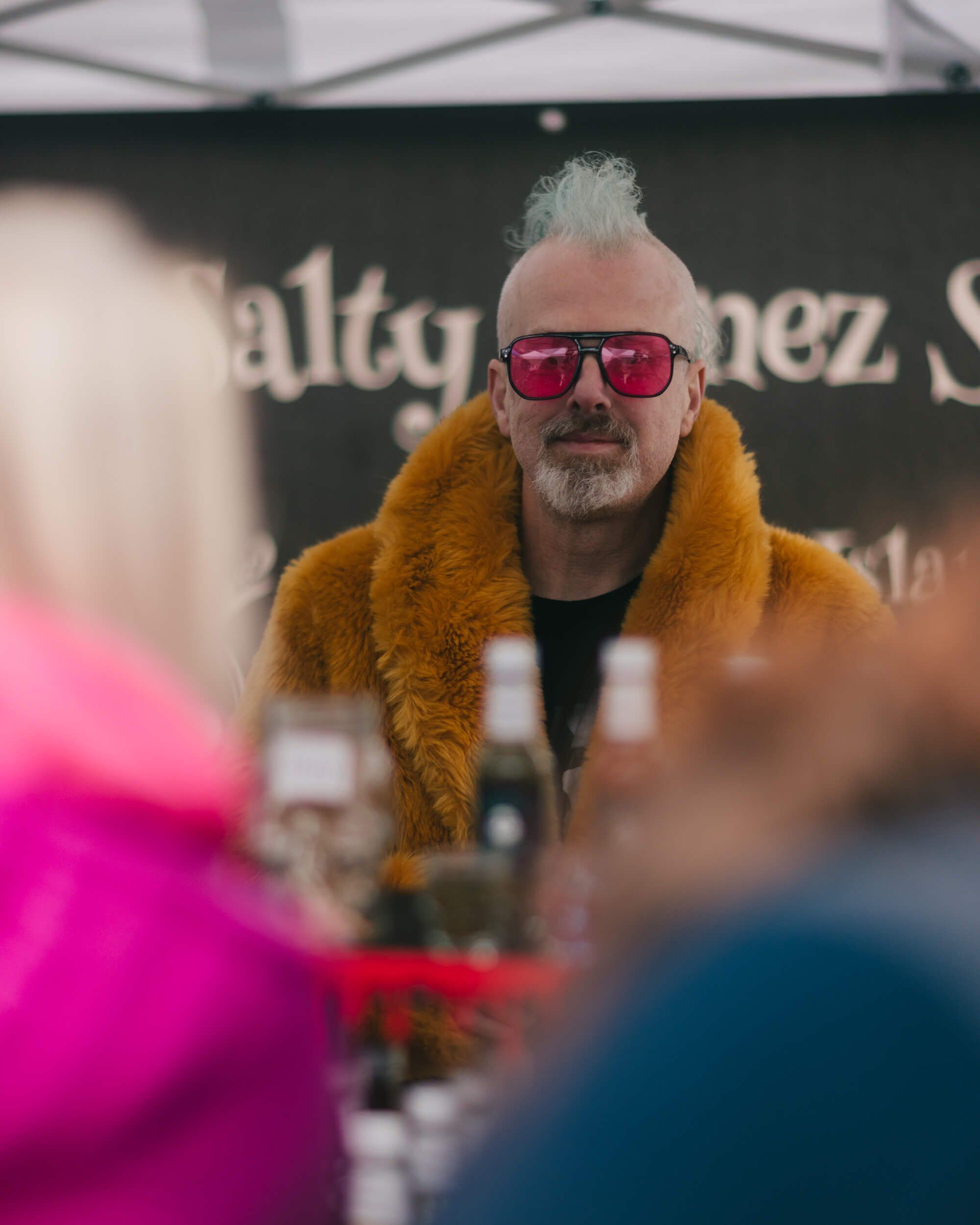 Man with white spiky hair and pink sunglasses wearing a mustard-yellow fur collar smiles at Saturday Market among blurred vendors and shoppers