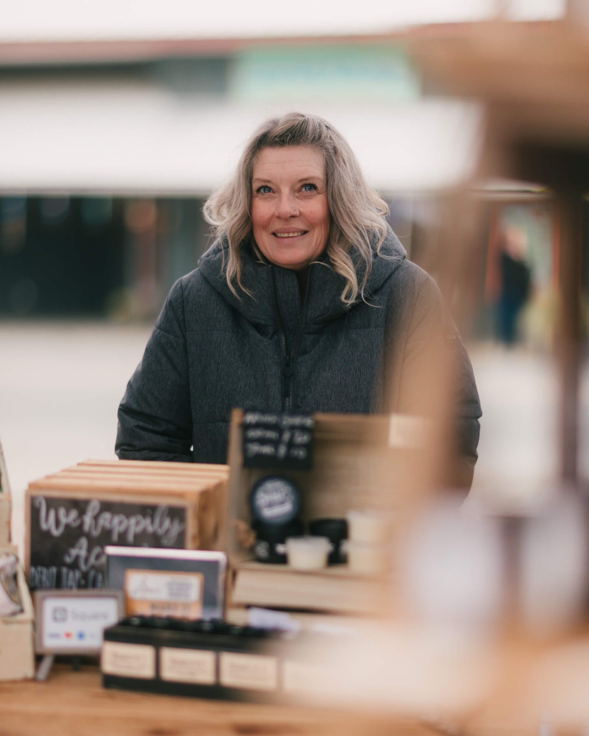 Woman with long gray hair smiles behind market vendor table with displayed products in soft natural light