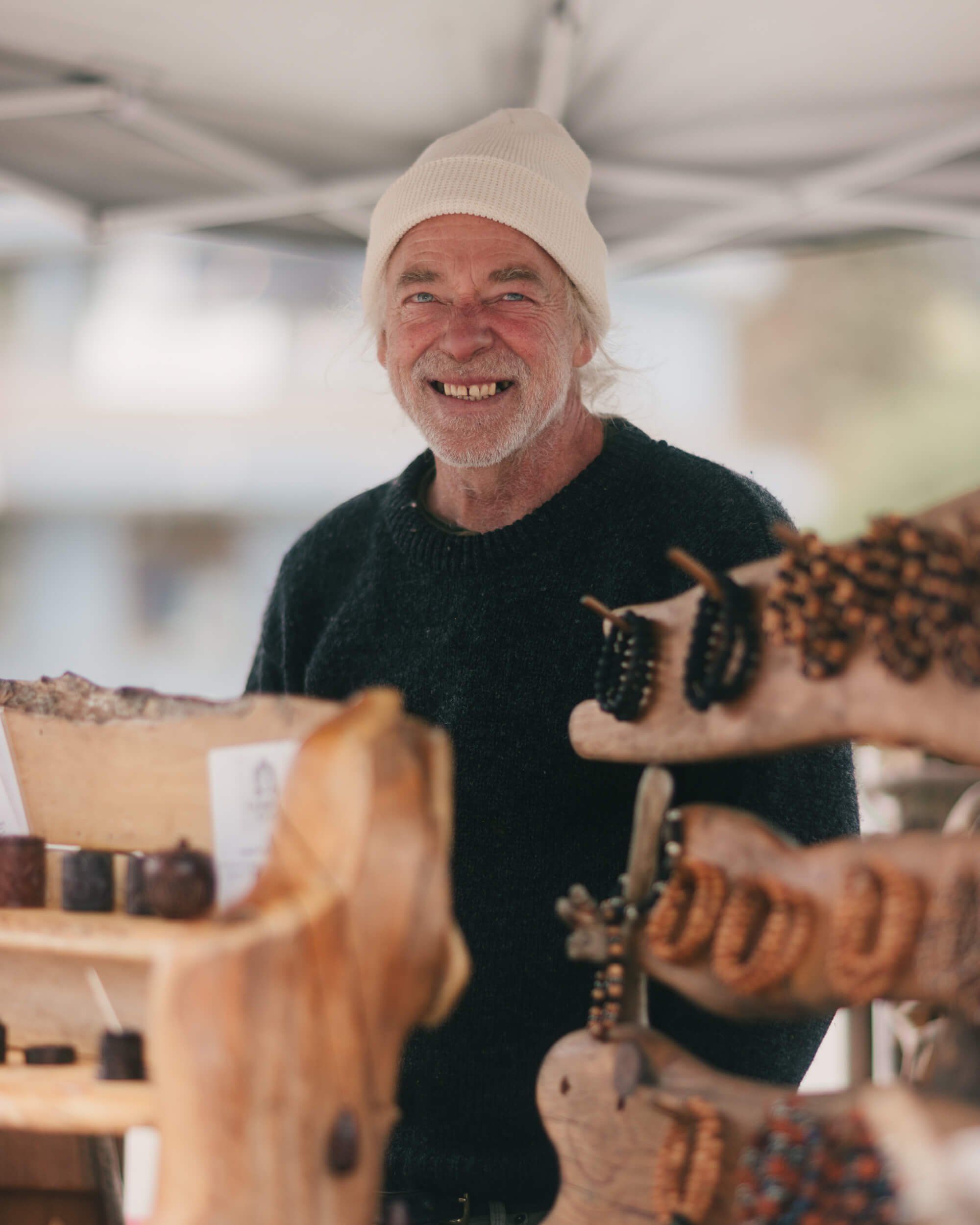 Smiling craftsperson in cream beanie displays wooden guitar bodies at an indoor market stall