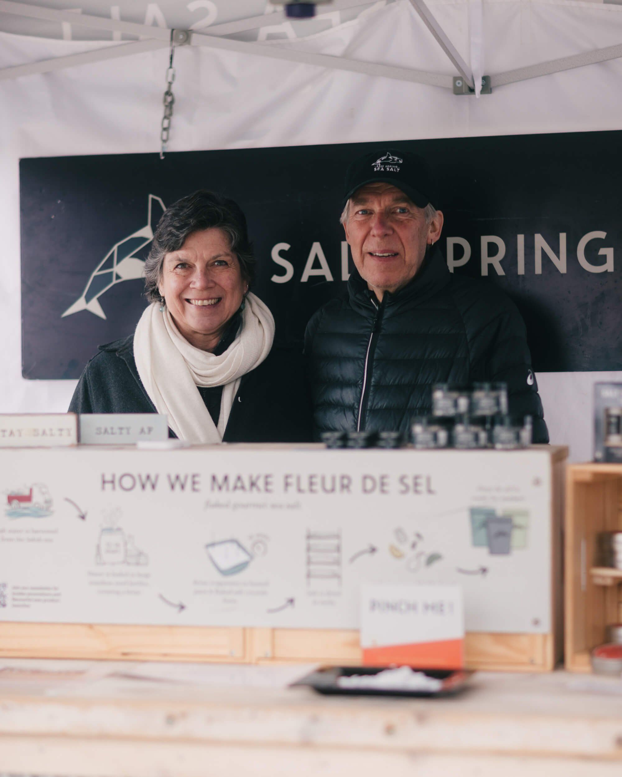 Two vendors smile behind a wooden market booth displaying salt production infographics under a white tent canopy