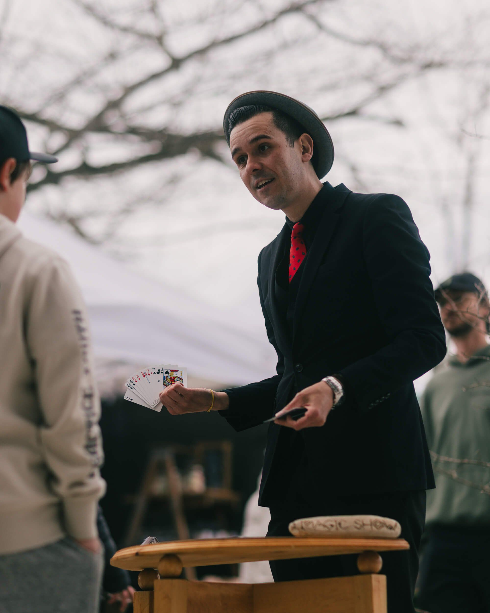 Man in black blazer and red shirt gestures while speaking at outdoor market table, bare branches visible overhead