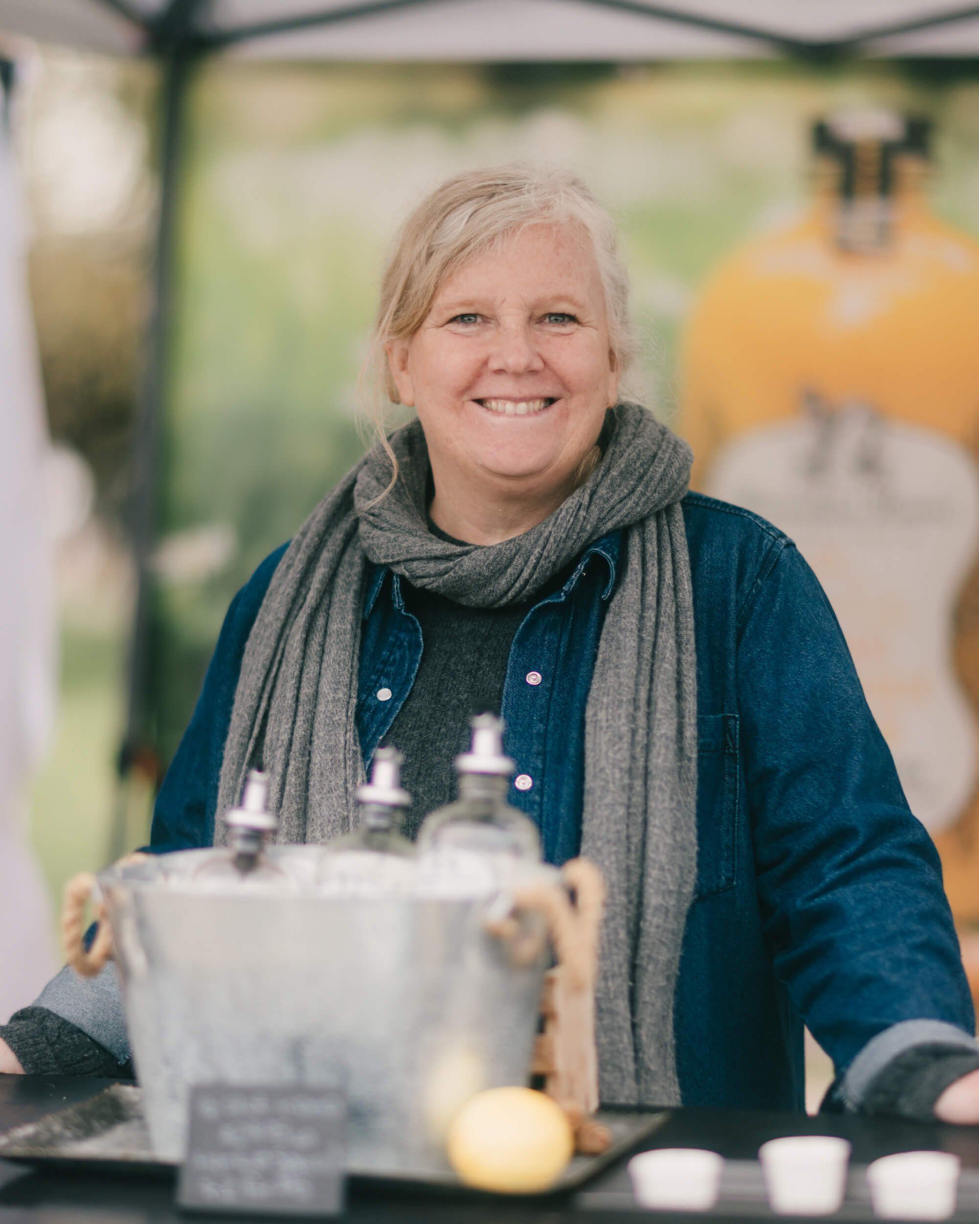 Smiling vendor at outdoor market stand with glass bottles and produce, wearing navy shirt and gray scarf on sunny day