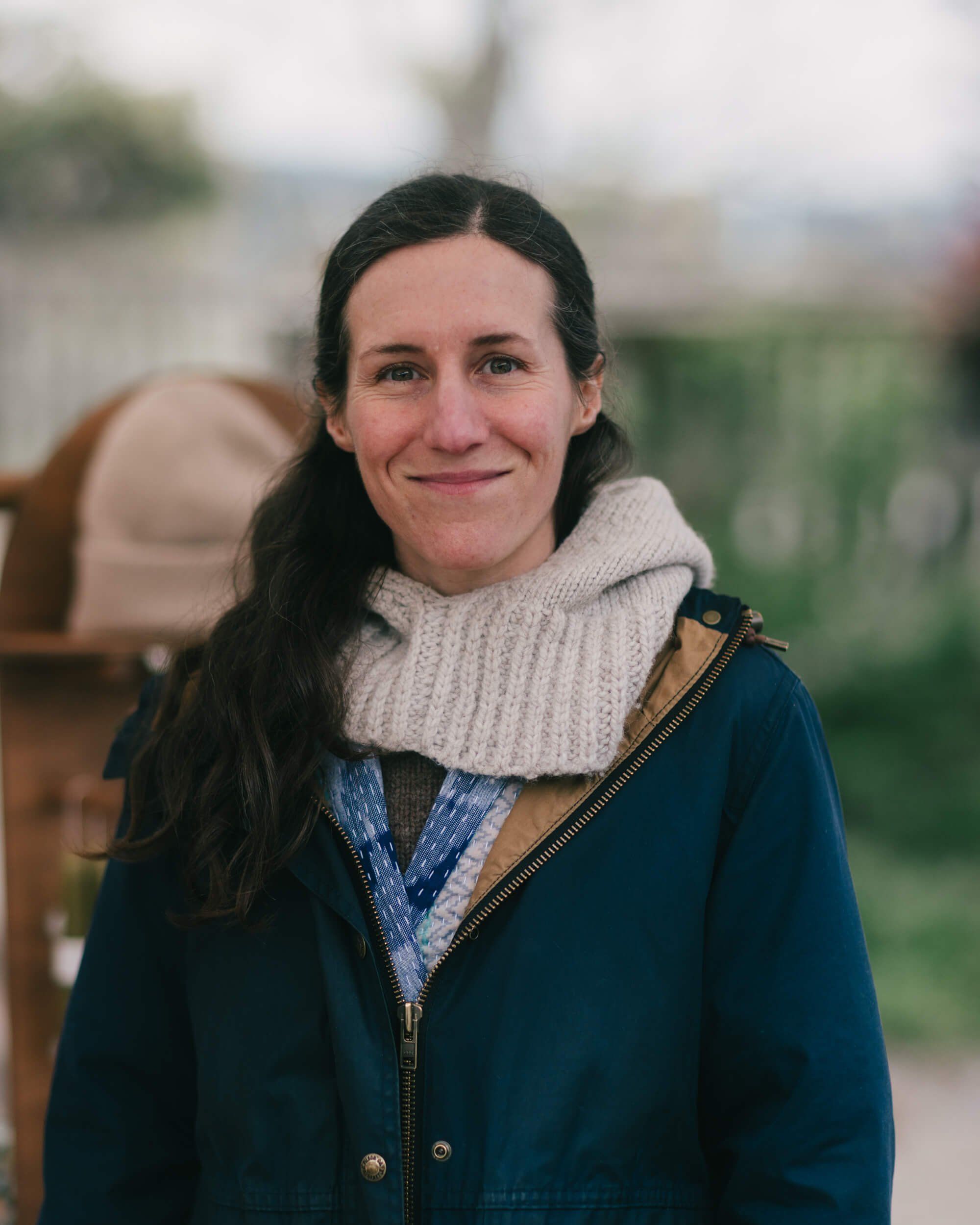 Woman with long dark hair wearing a cream cowl neck and navy jacket smiles at the Saturday Market, soft natural light and blurred green background