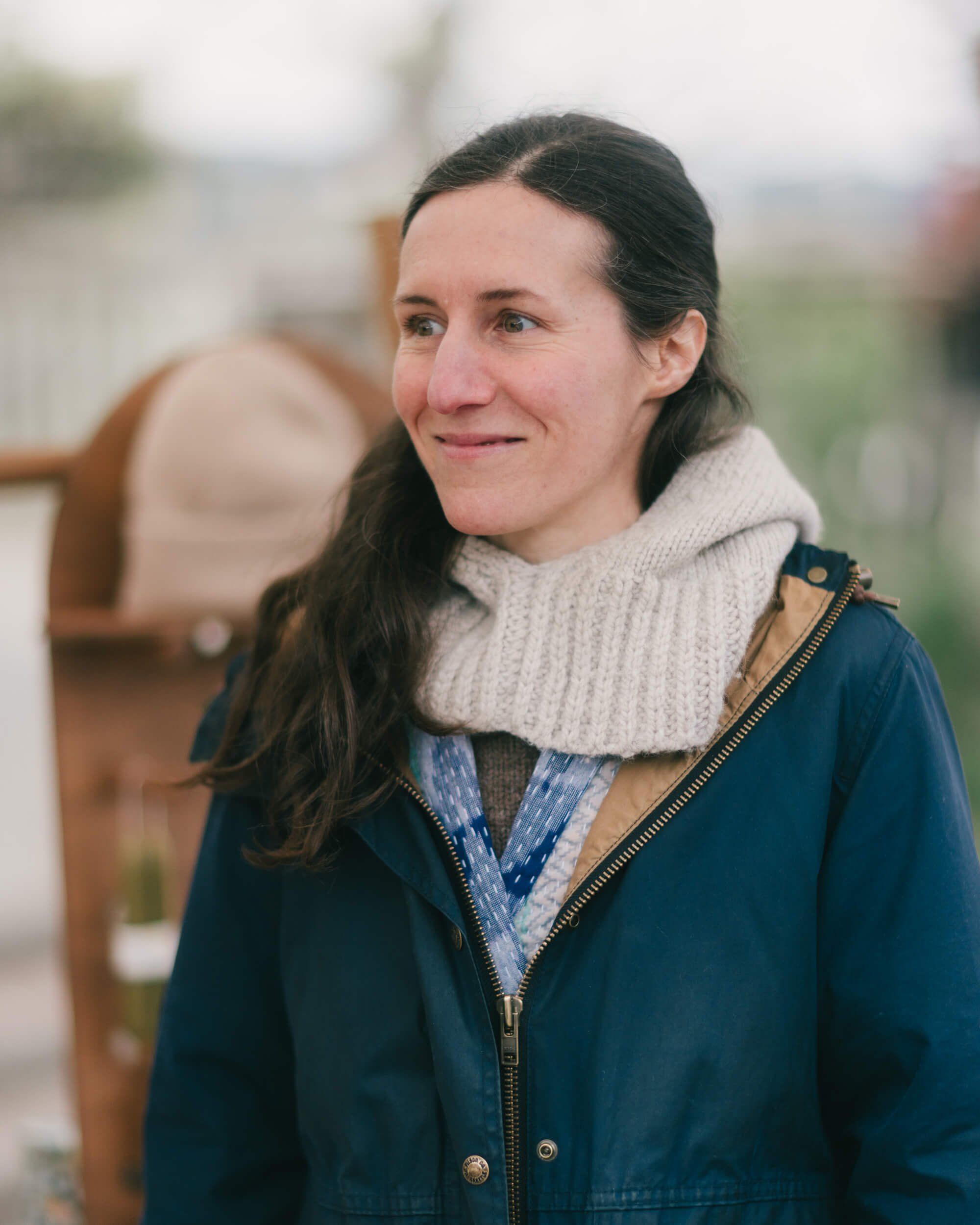 Woman with long dark hair wearing a navy jacket and cream knit cowl neck, smiling warmly outdoors with soft natural light