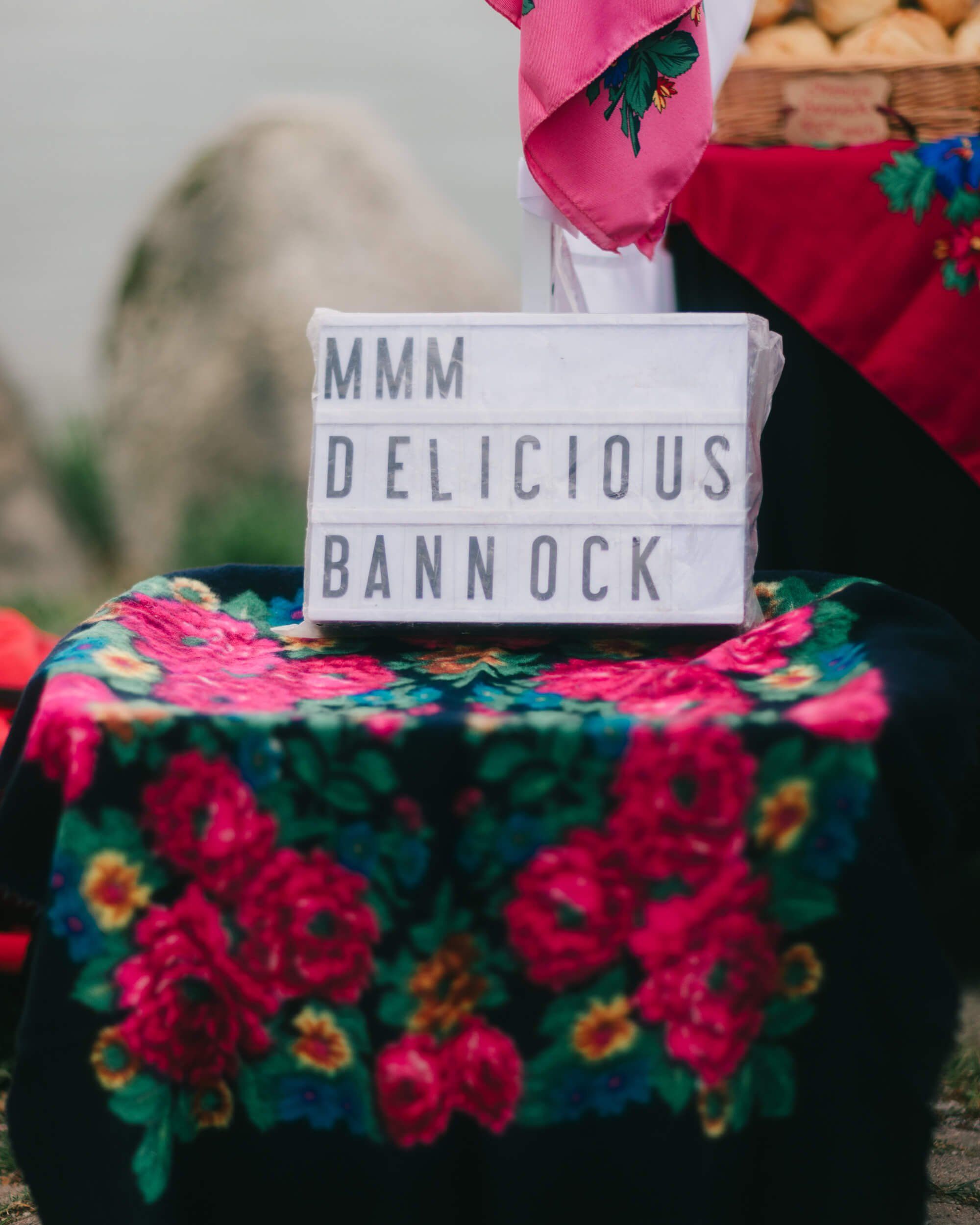 Handwritten sign reading "MMM DELICIOUS BANNOCK" sits atop vibrant floral fabric at an outdoor market stall