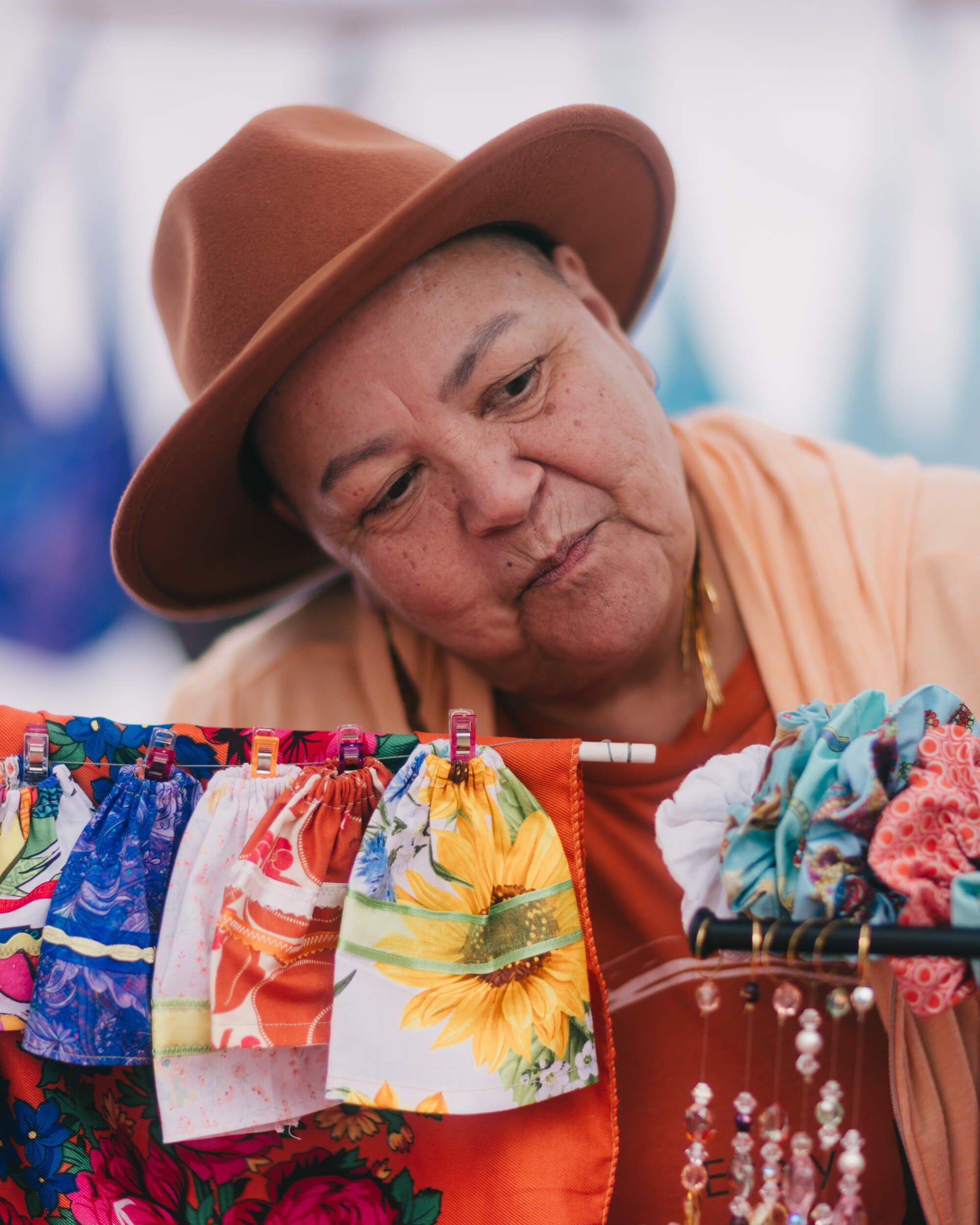 Vendor in terracotta hat smiles warmly while displaying colorful patterned fabrics and textiles at market stall