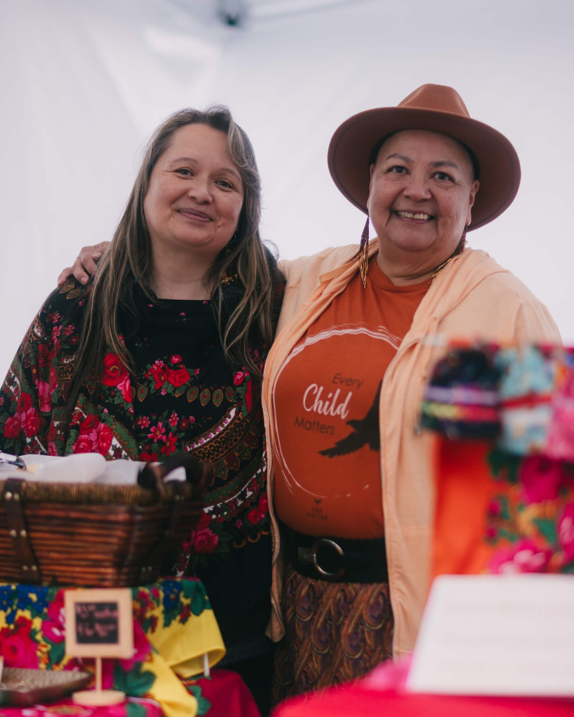 Two women smile warmly at Saturday Market, one wearing a floral jacket and the other in an orange cardigan and cowboy hat, with colorful crafts displayed in…