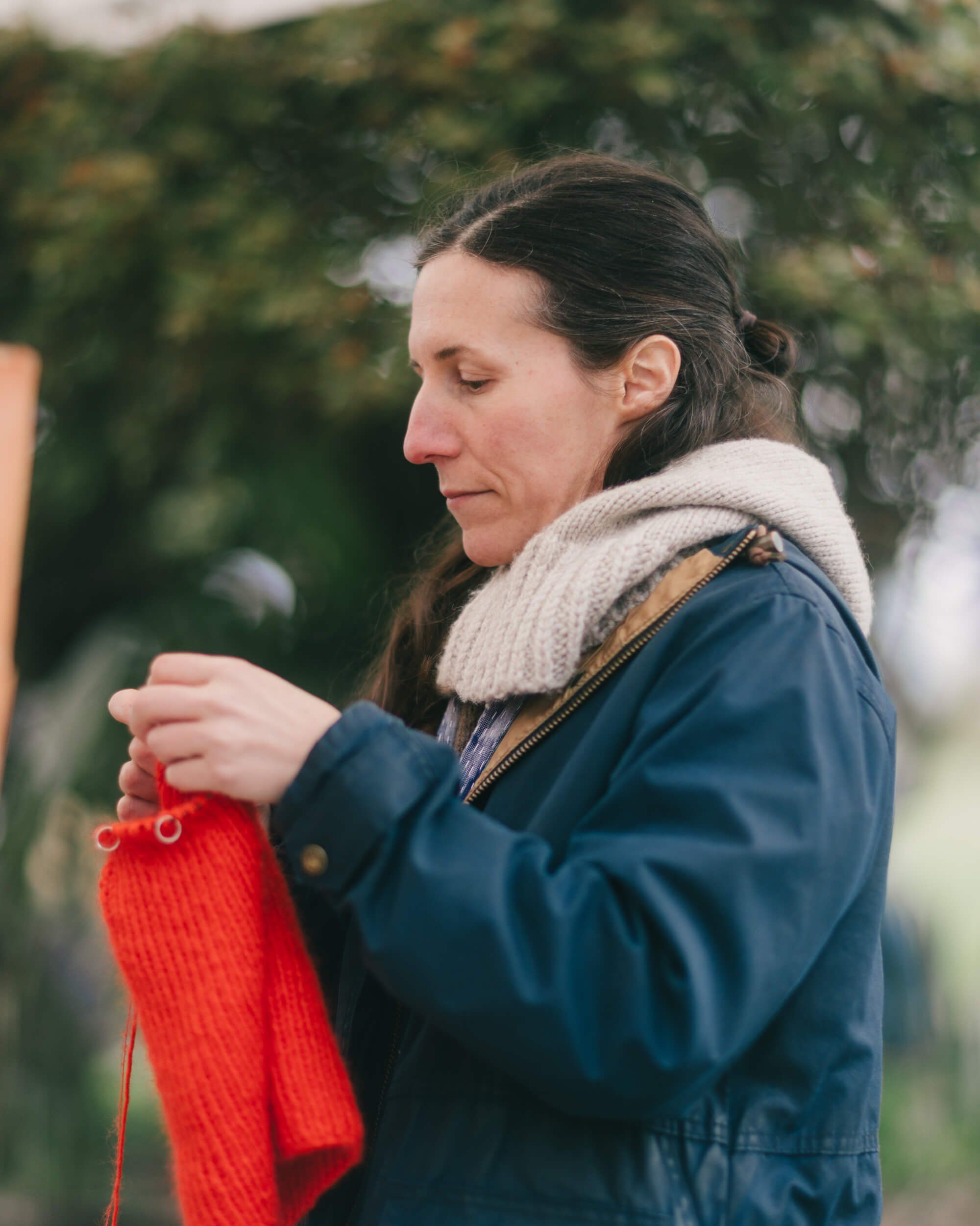 Woman in blue jacket holding a red knitted garment at an outdoor market, soft natural light filtering through trees behind her
