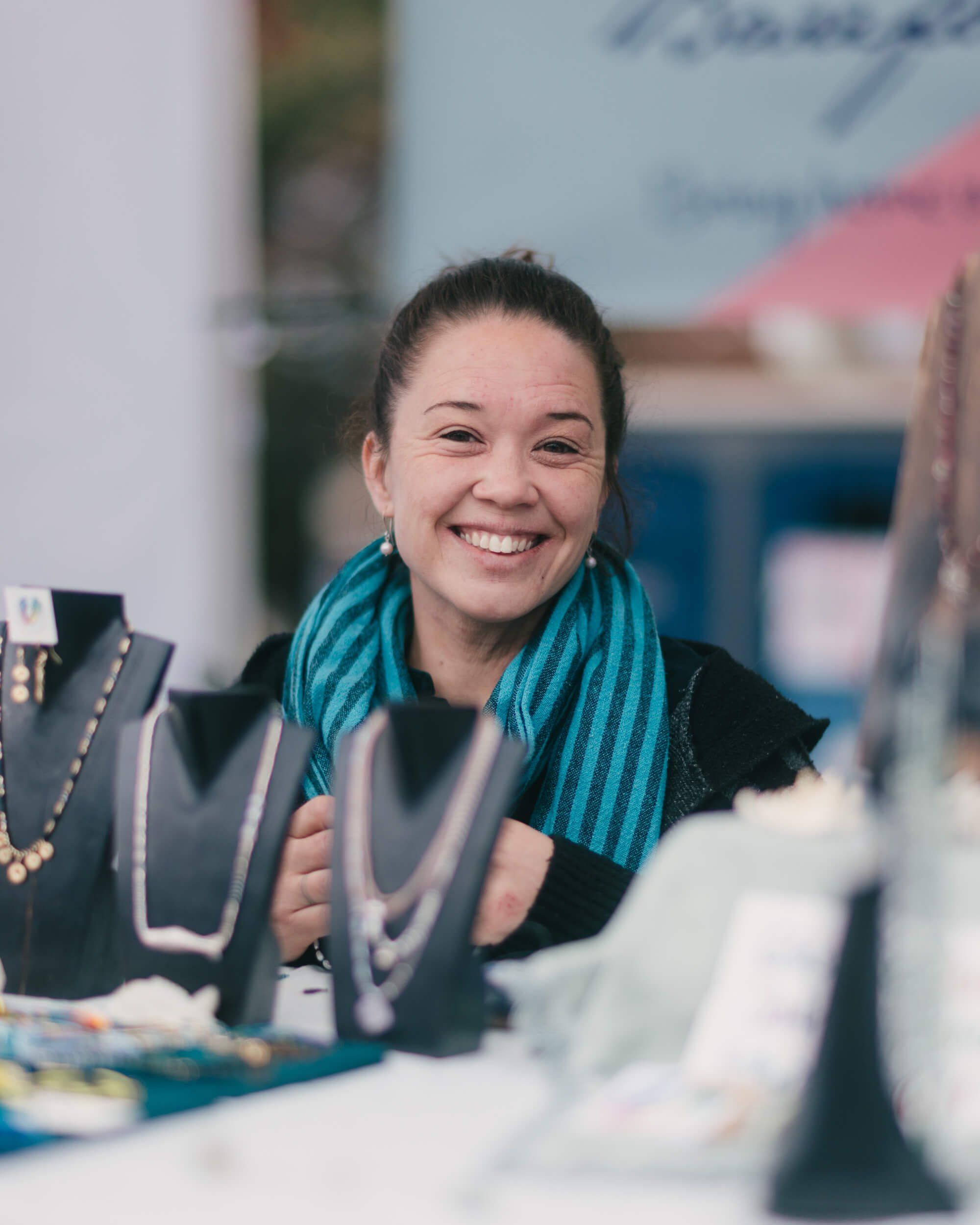Smiling vendor displays jewelry at a market stall, wearing a teal striped scarf and black top
