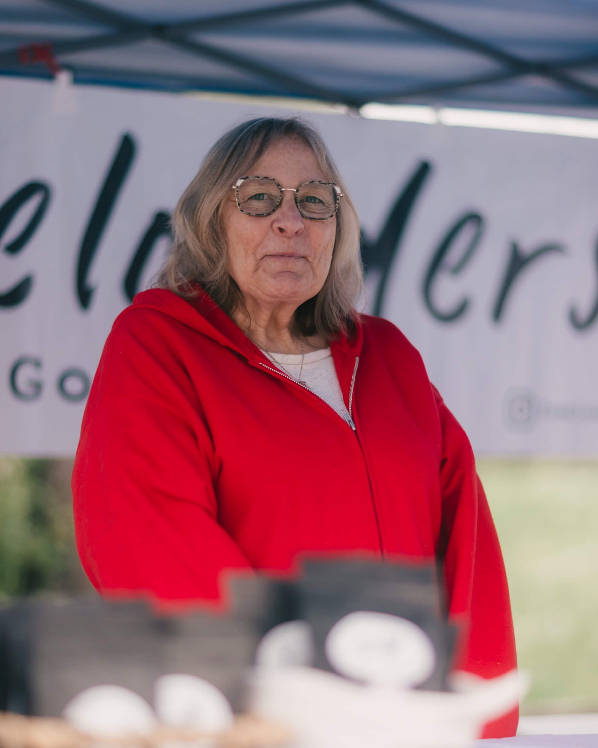 Woman in red jacket and glasses at farmers market stand under tent, with eggs visible in foreground