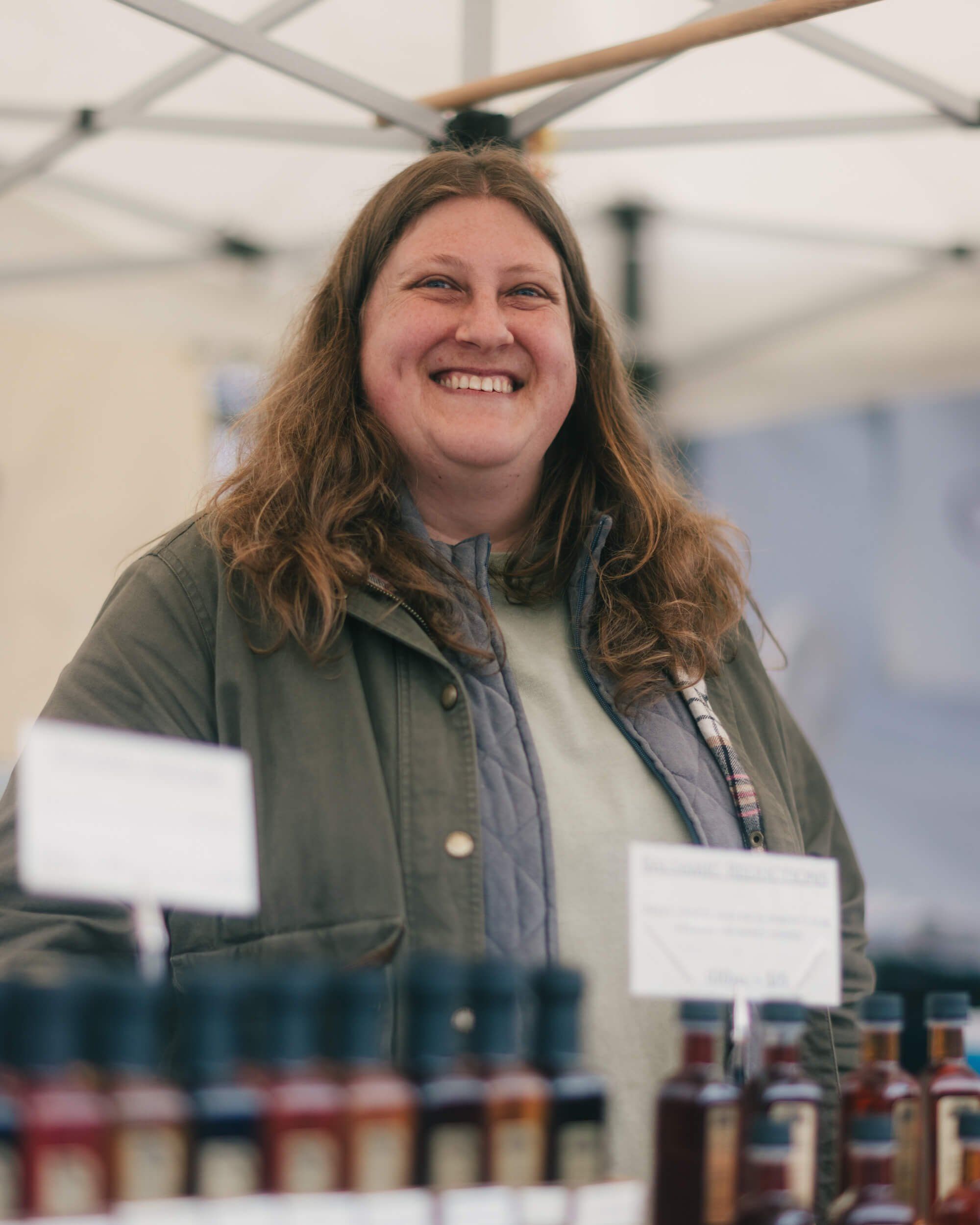 Smiling vendor at farmers market tent with glass jars displayed on table below