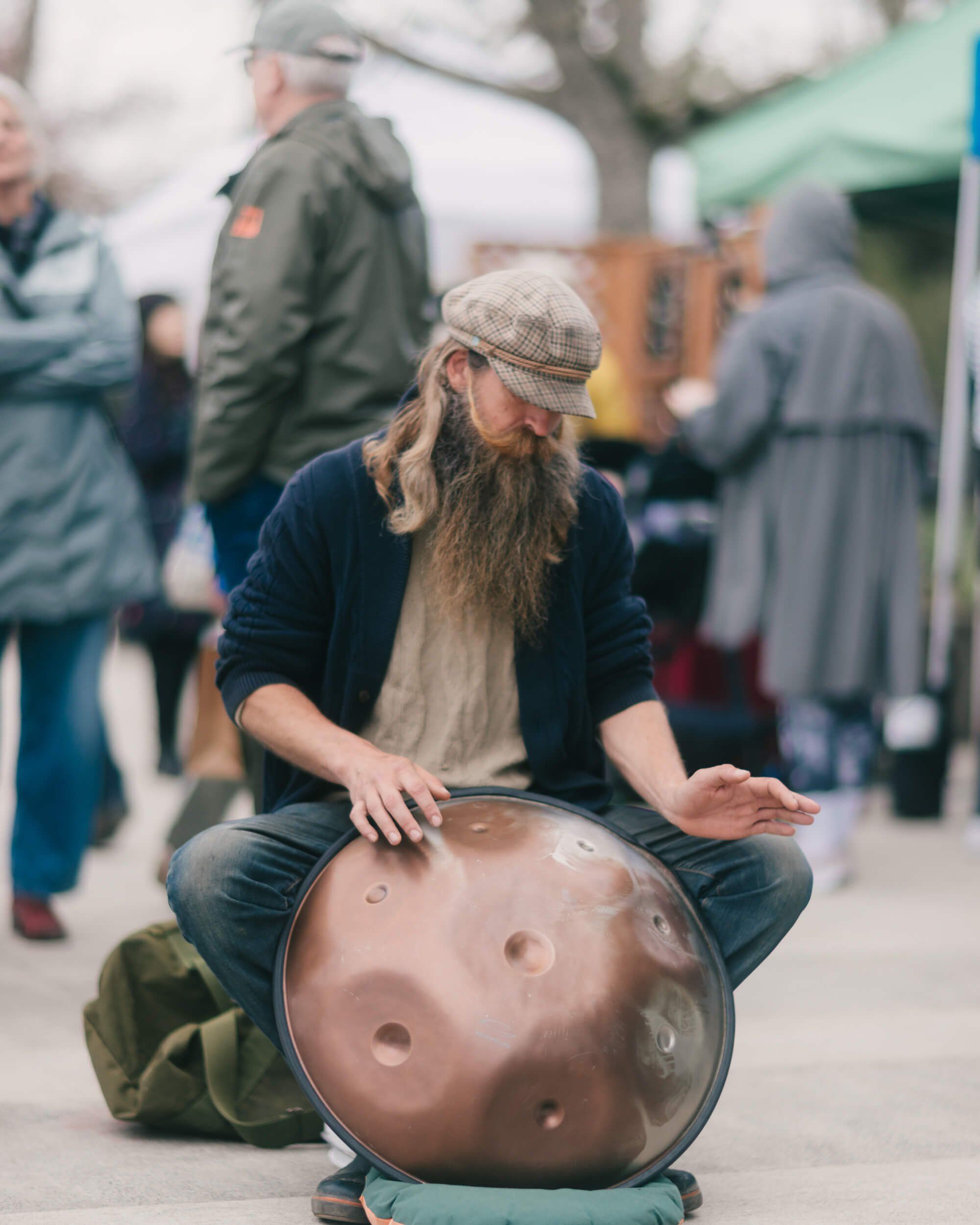 Bearded musician playing a handpan drum at an outdoor market, surrounded by visitors on a overcast day