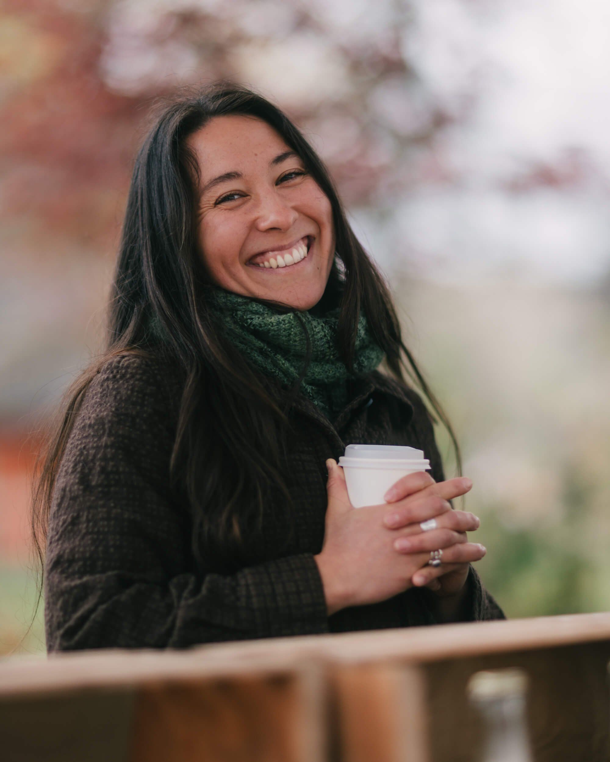 Smiling woman with long dark hair holding a white cup, wearing a dark sweater and green scarf at an outdoor market