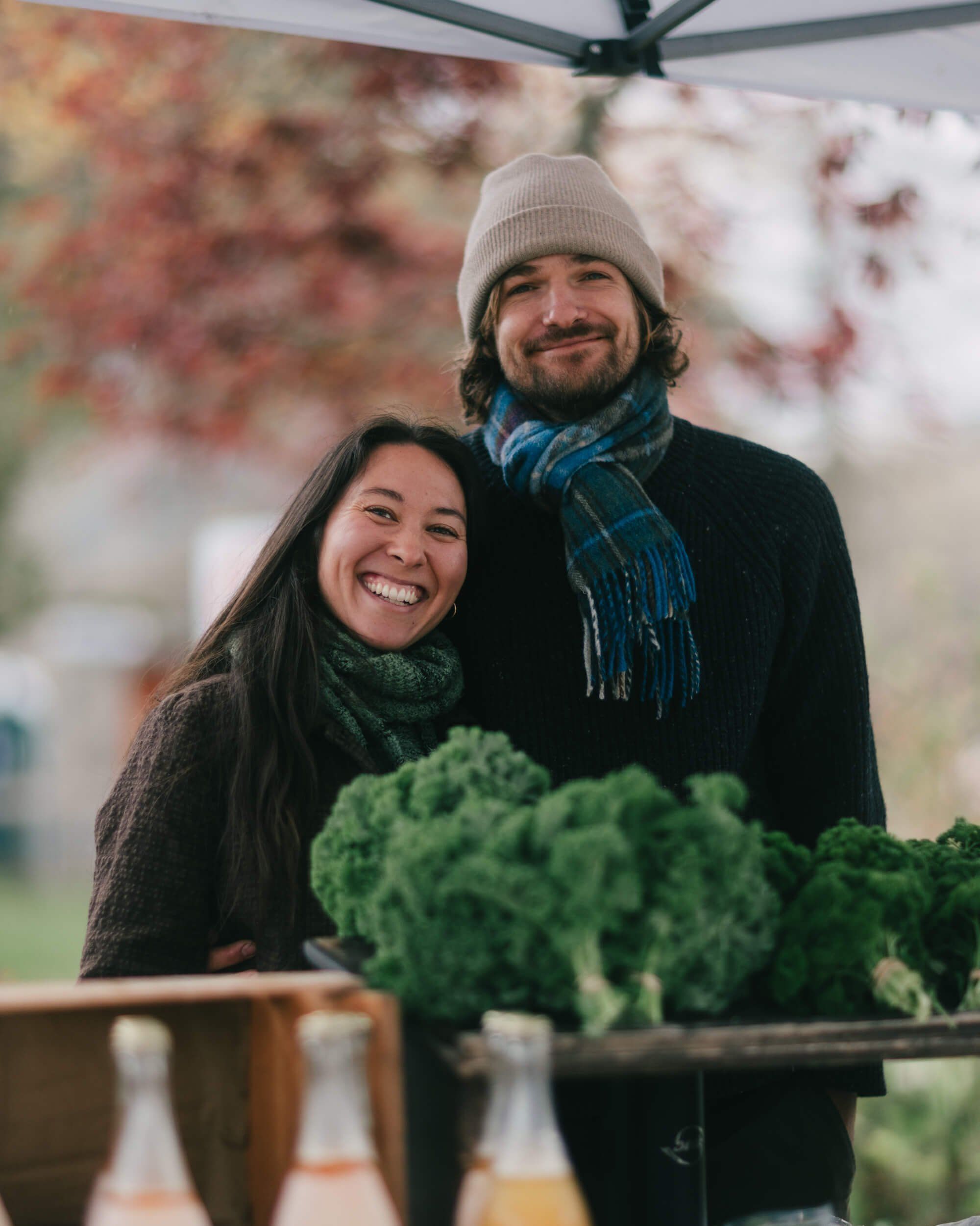 Couple smiling under market tent with fresh kale, wearing winter layers on a cool autumn day