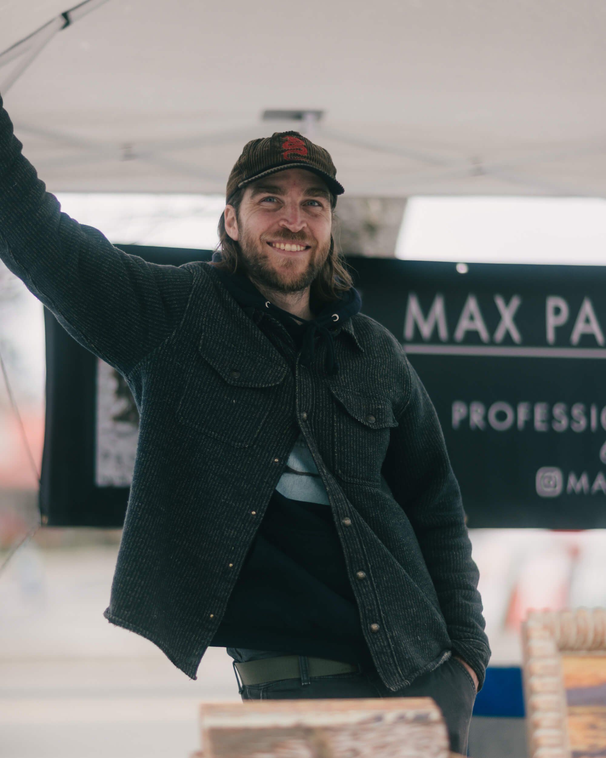 Bearded man in black denim jacket and cap waving under a market tent, smiling at the camera