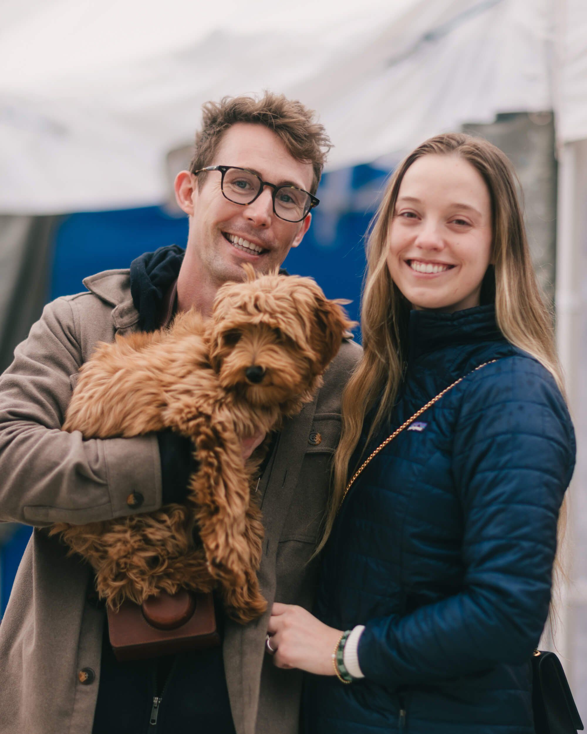 Couple holding a golden doodle puppy, smiling warmly at Saturday market with blue tent backdrop