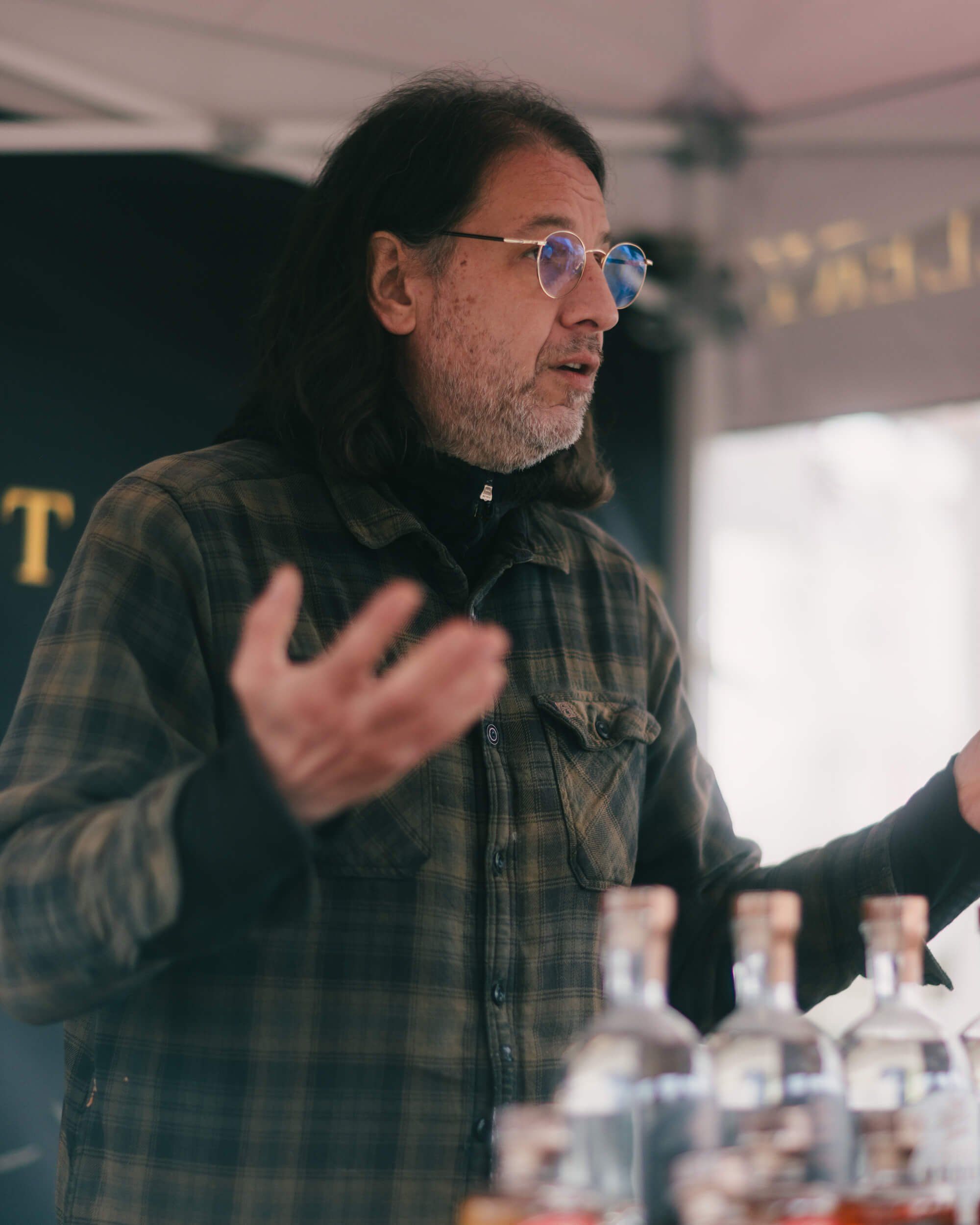 Man in plaid shirt and glasses gestures expressively while speaking at an indoor market event, with water glasses visible on the table
