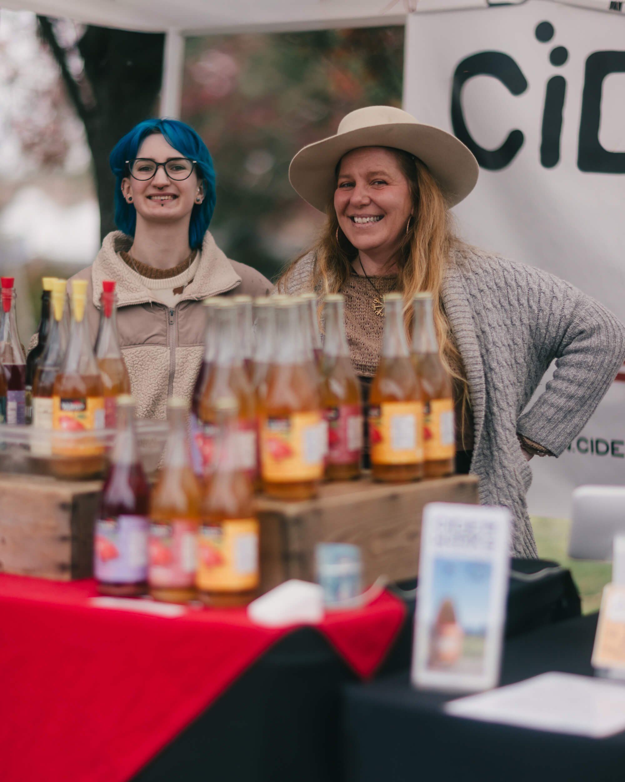 Two smiling vendors at a market booth display colorful glass jars with candles under a red-draped table and signage