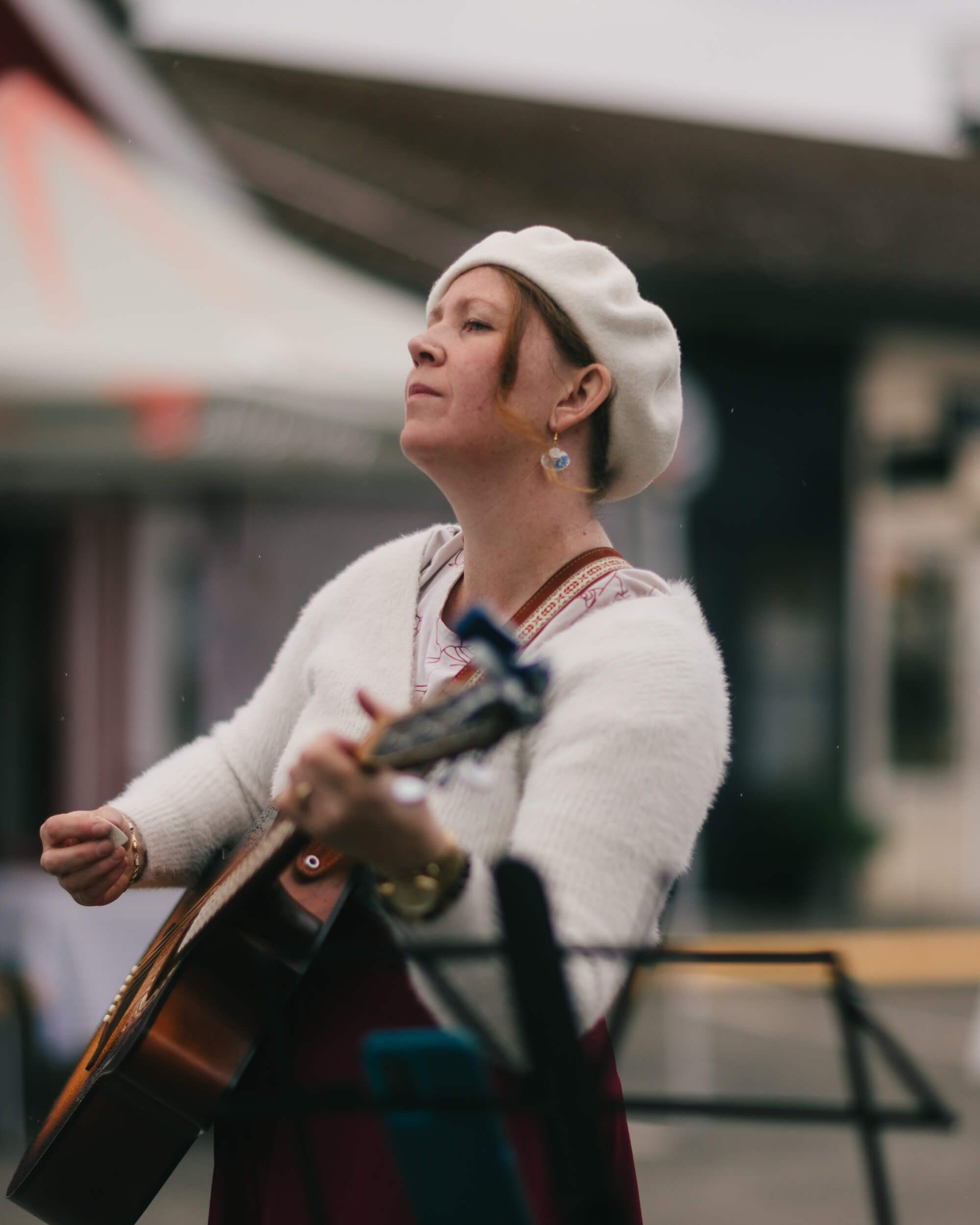 Woman in cream beret and white cardigan plays guitar at outdoor market, looking upward with soft natural light