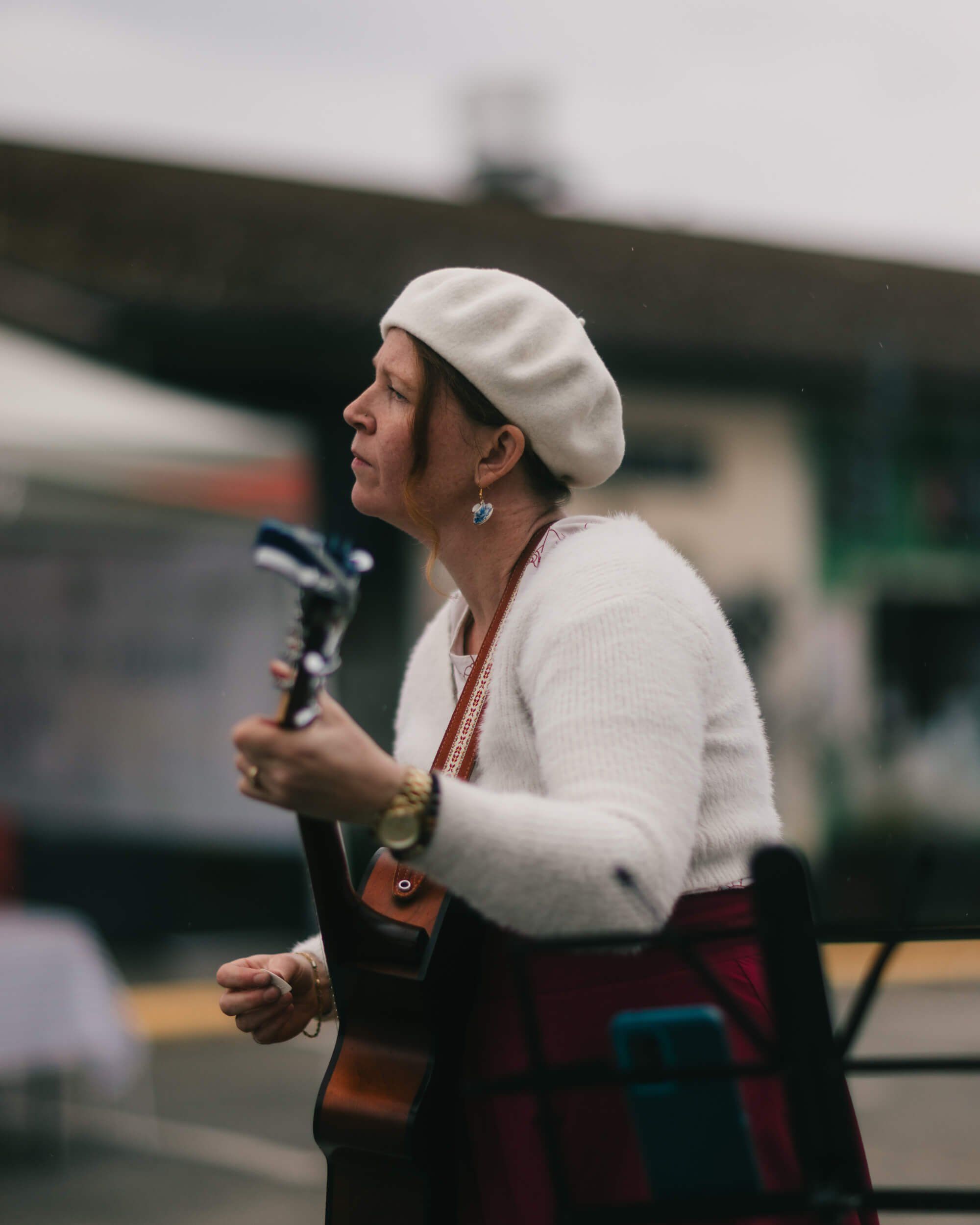 Woman in white beret and burgundy skirt speaking at microphone at outdoor market on overcast day