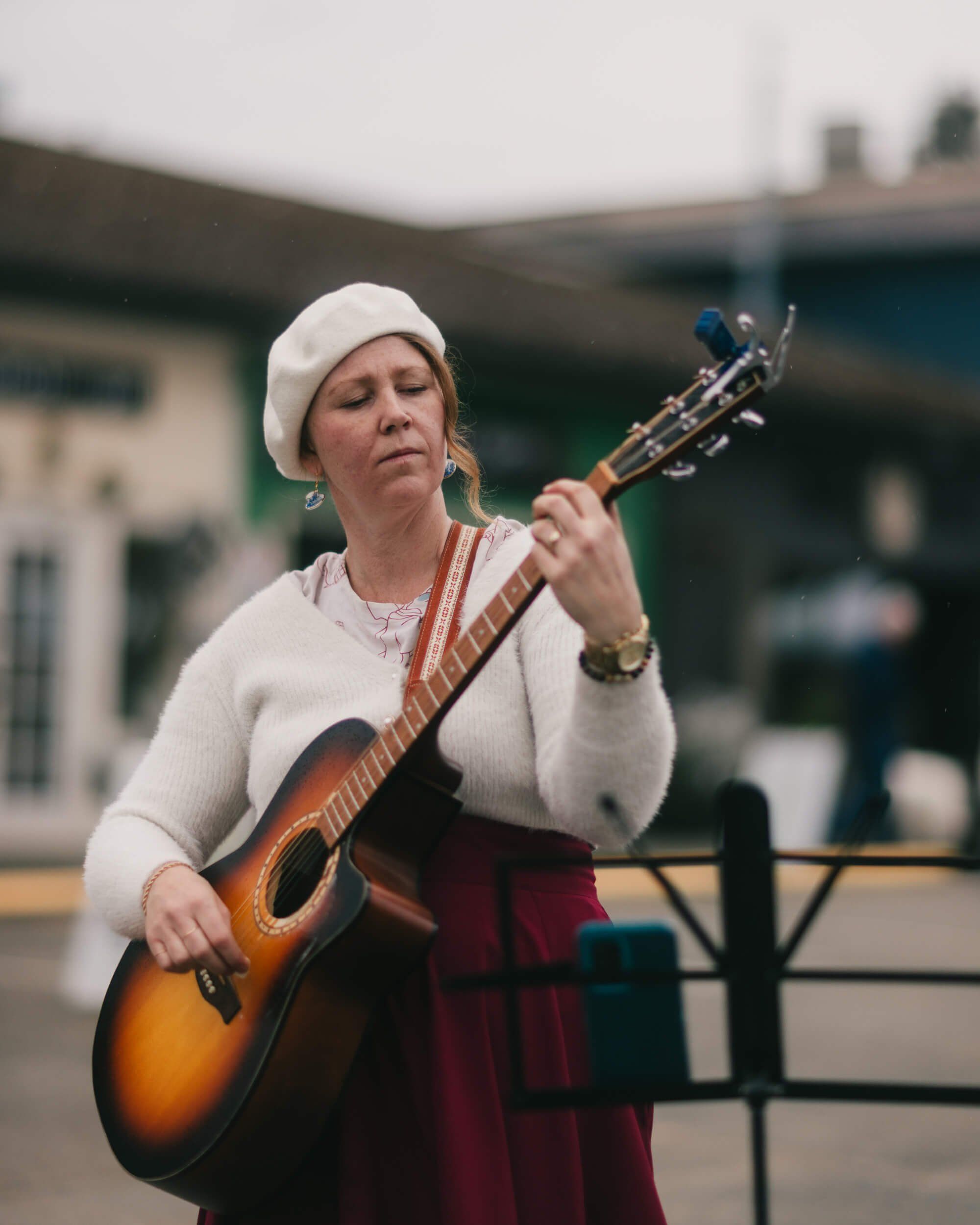 Musician in cream sweater and white beanie plays acoustic guitar at outdoor market, soft daylight illuminating her focused expression against blurred buildin…