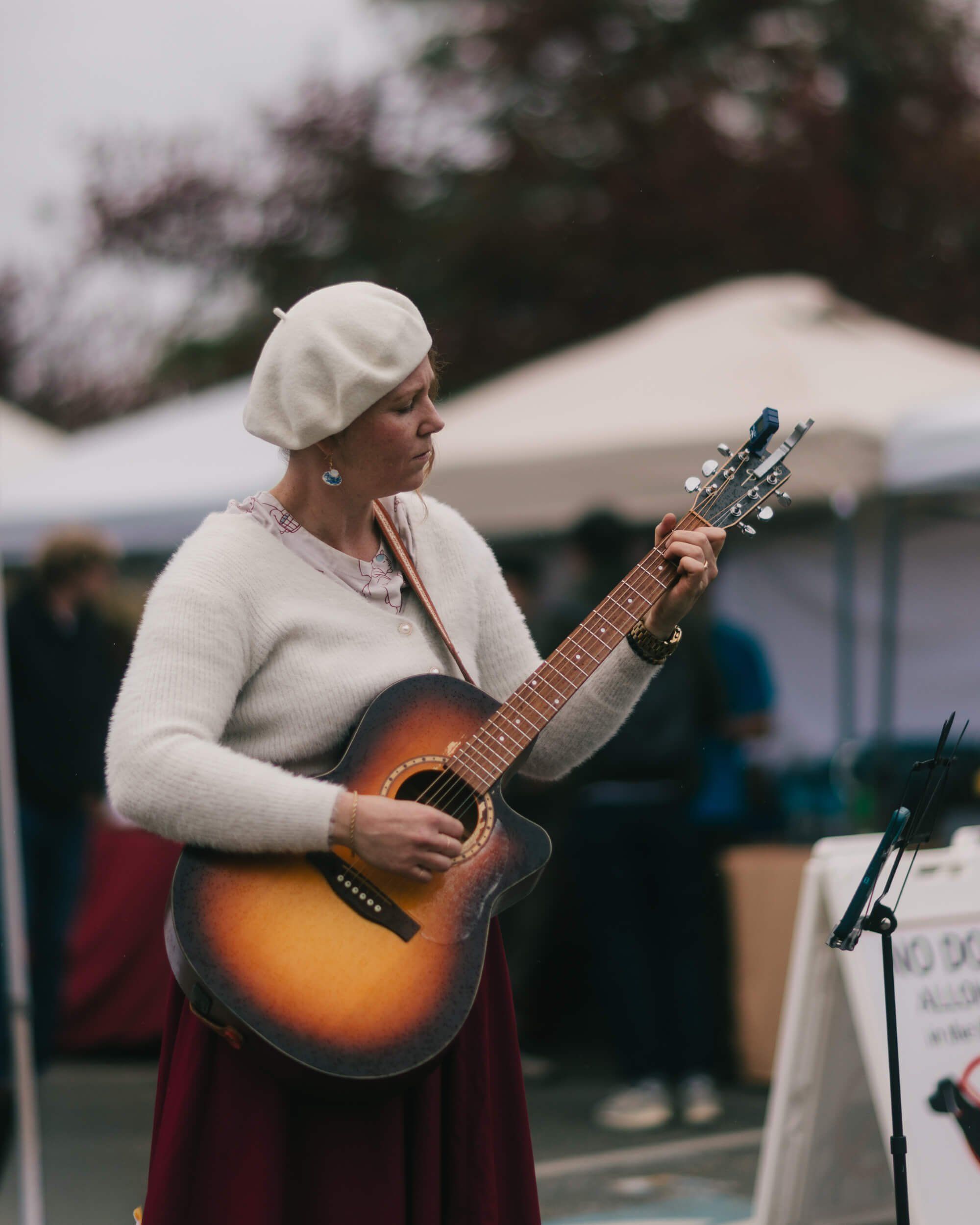 Woman in cream sweater and beret plays acoustic guitar at outdoor market under soft overcast light