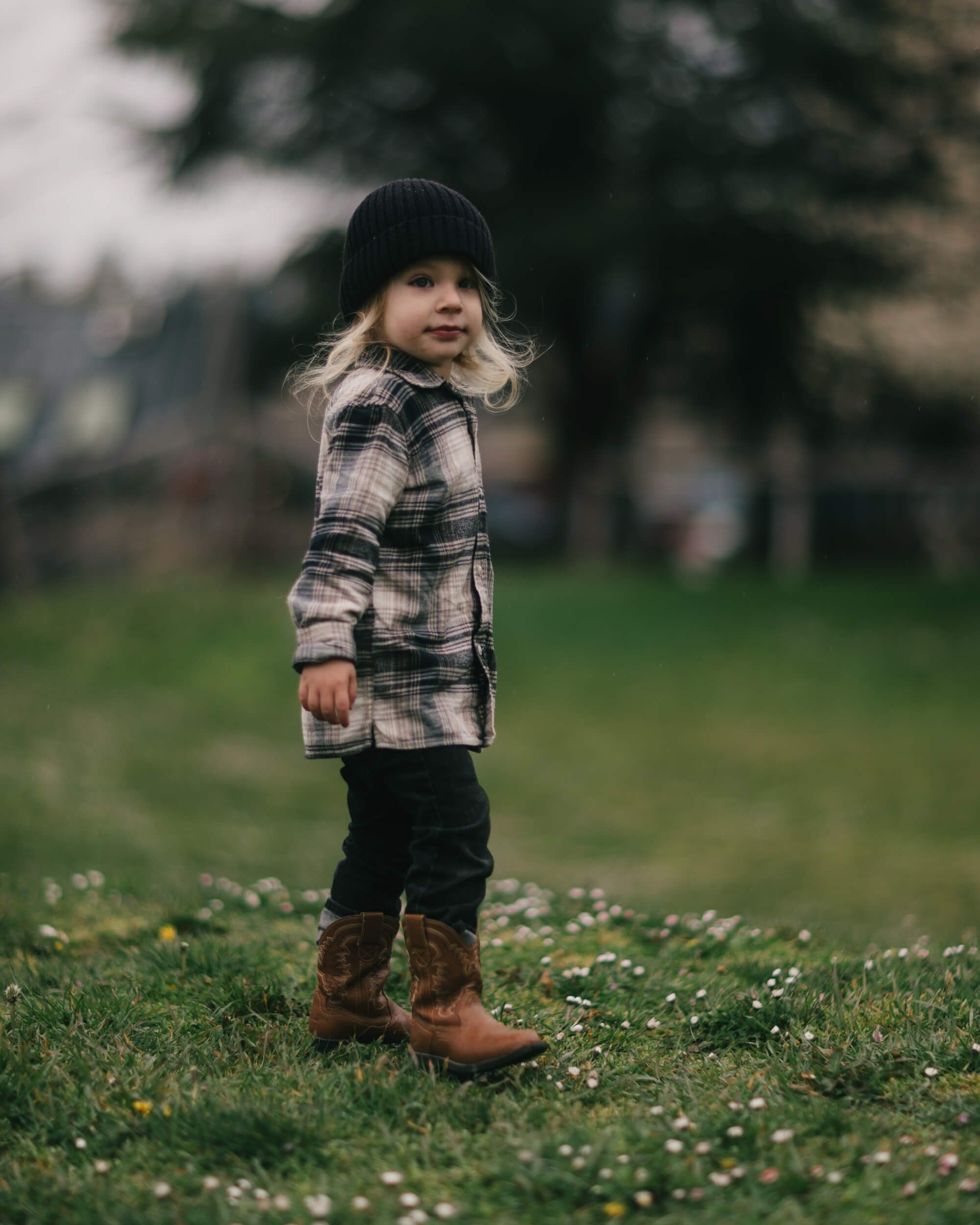 Toddler in plaid shirt and black beanie stands in grassy field scattered with wildflowers, looking back at camera with soft natural light