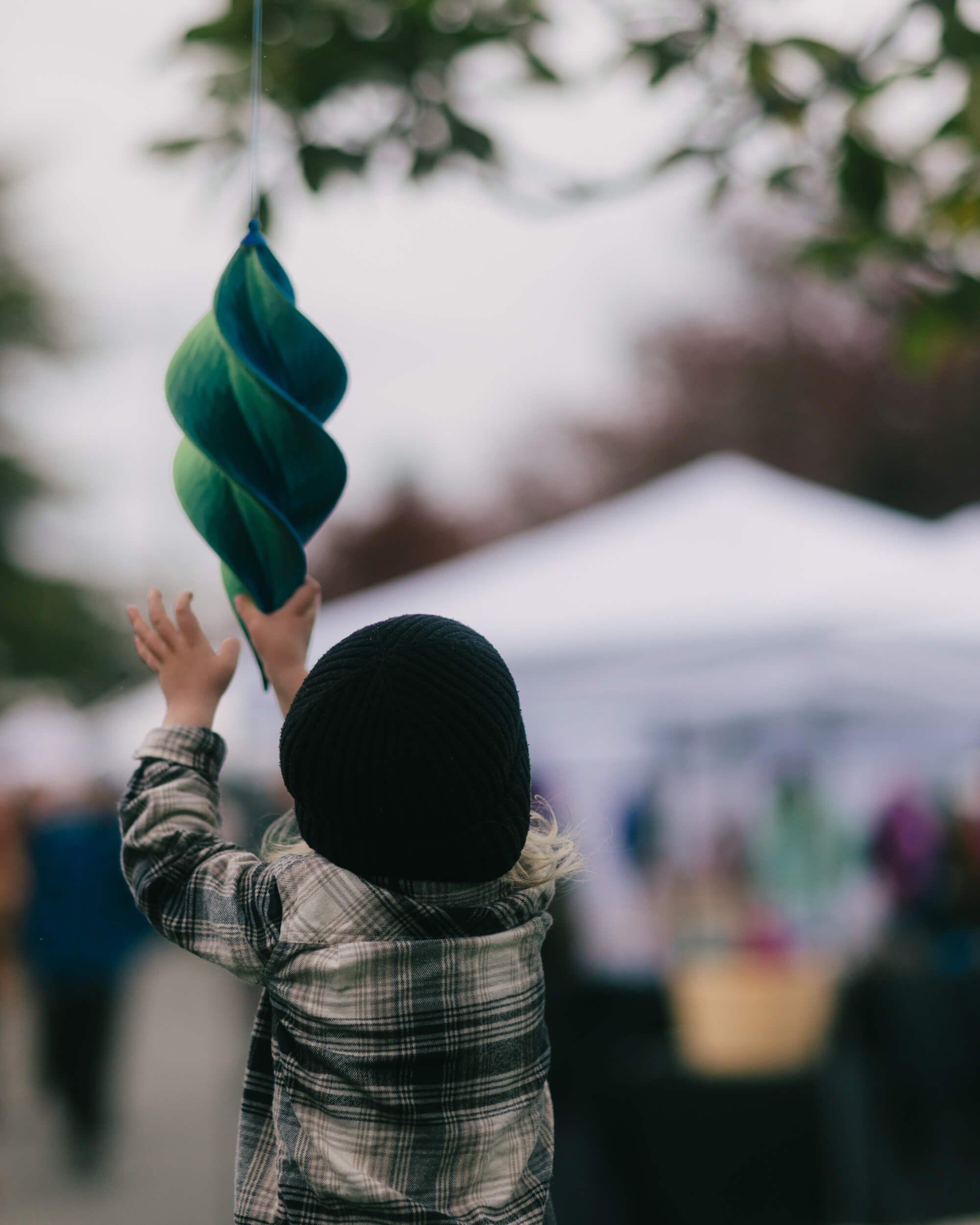 Child in black hat holding a twisted green leaf skyward at the Saturday Market, blurred vendors and tents behind