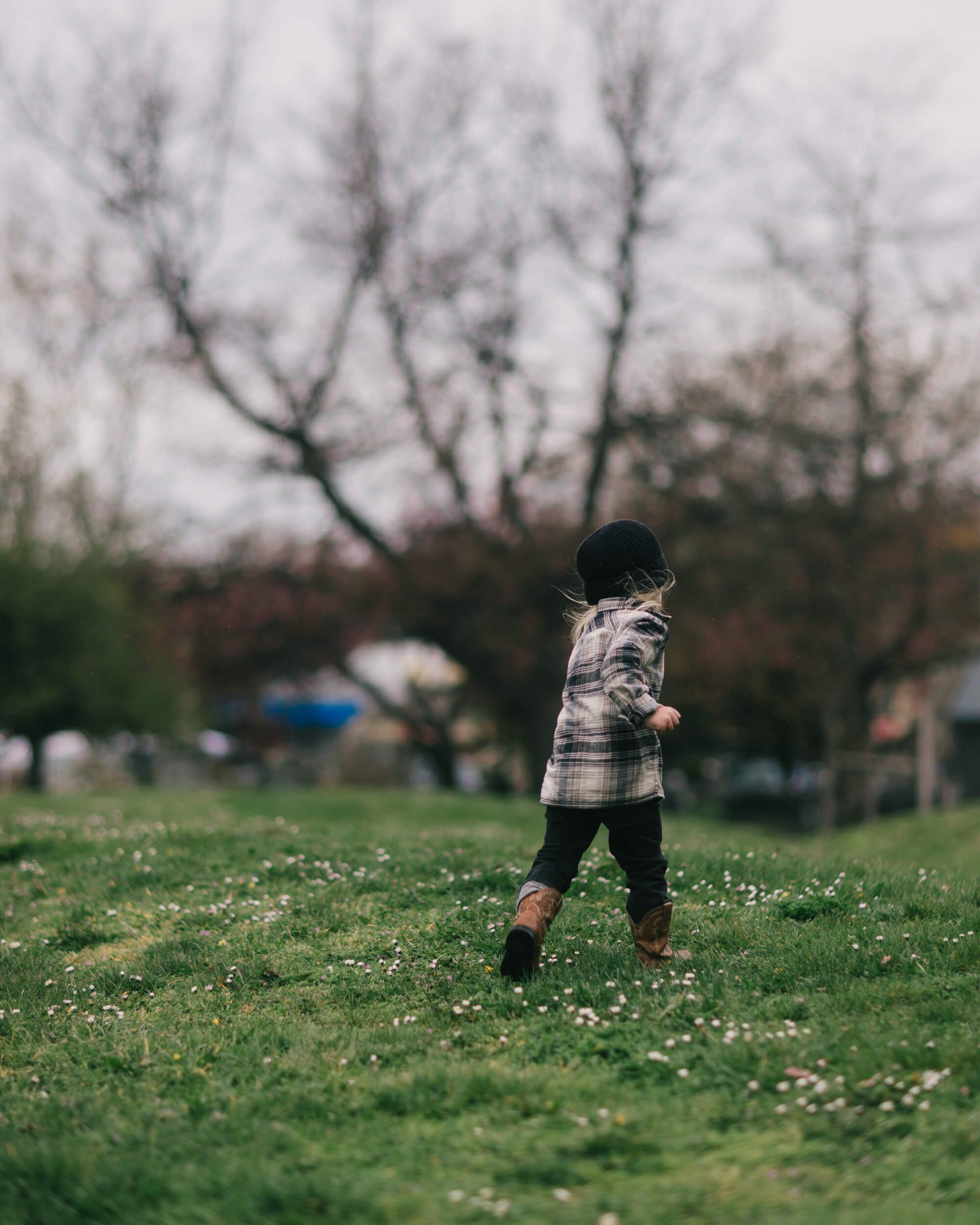 Toddler in plaid shirt running through grassy field with bare trees and soft overcast light