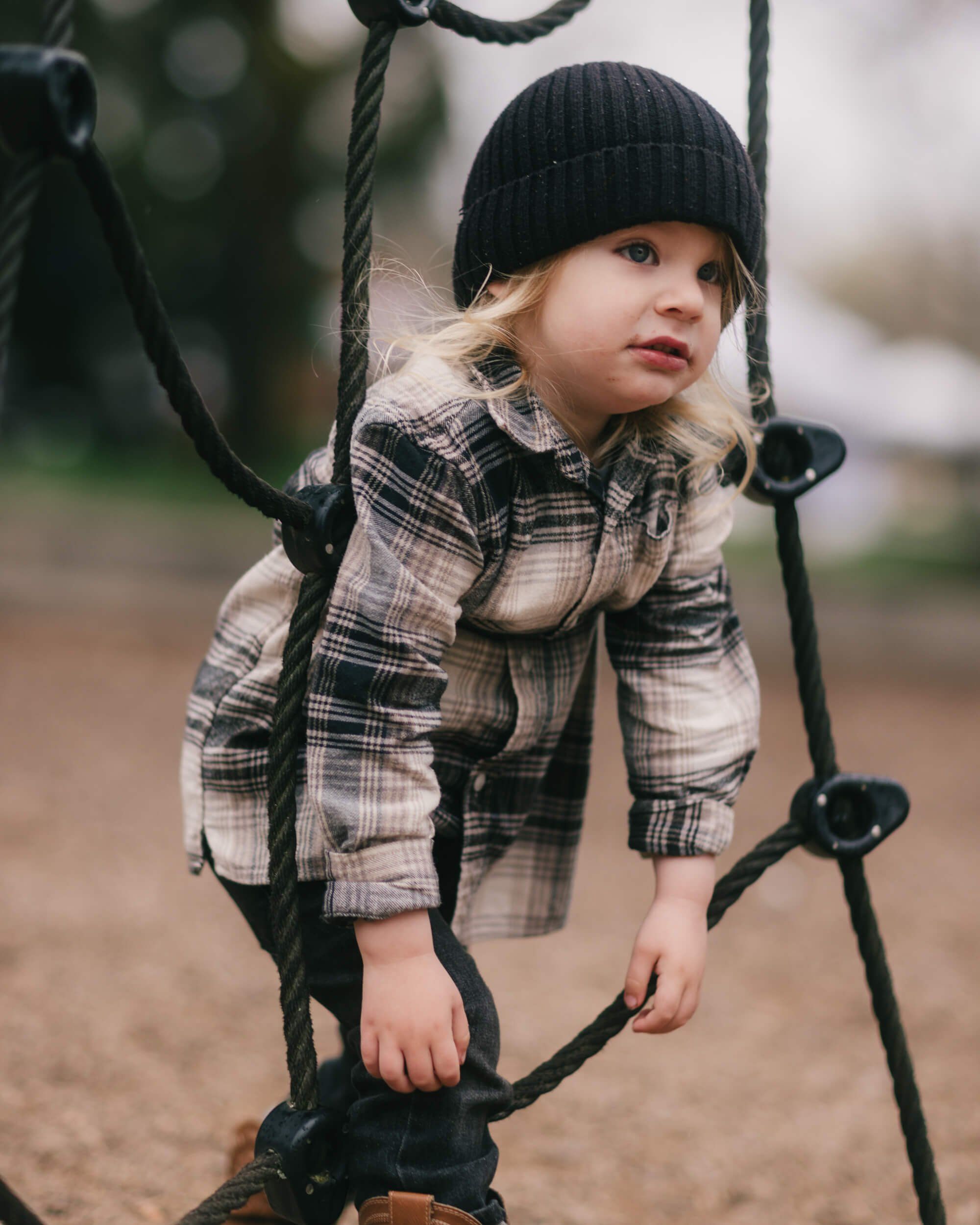 Young child in plaid shirt and black beanie climbing rope at playground, soft natural light