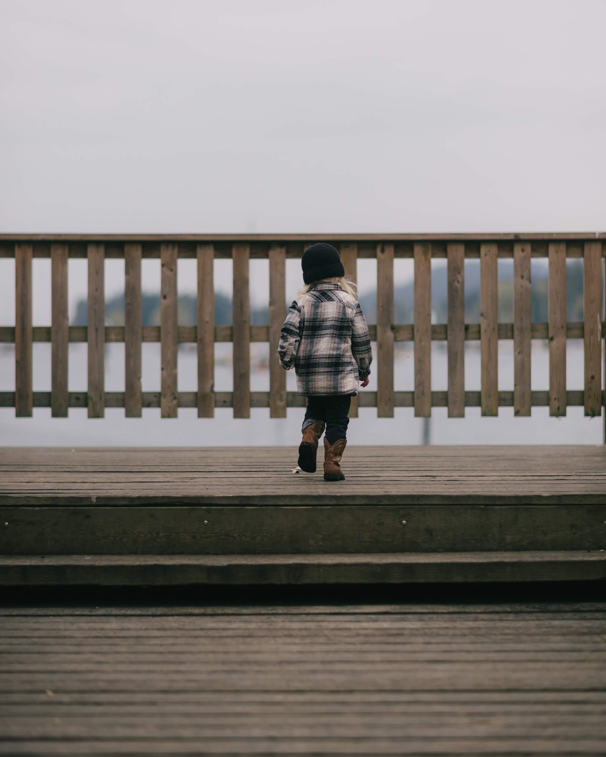 Child in plaid jacket running alone across misty wooden boardwalk toward white railing