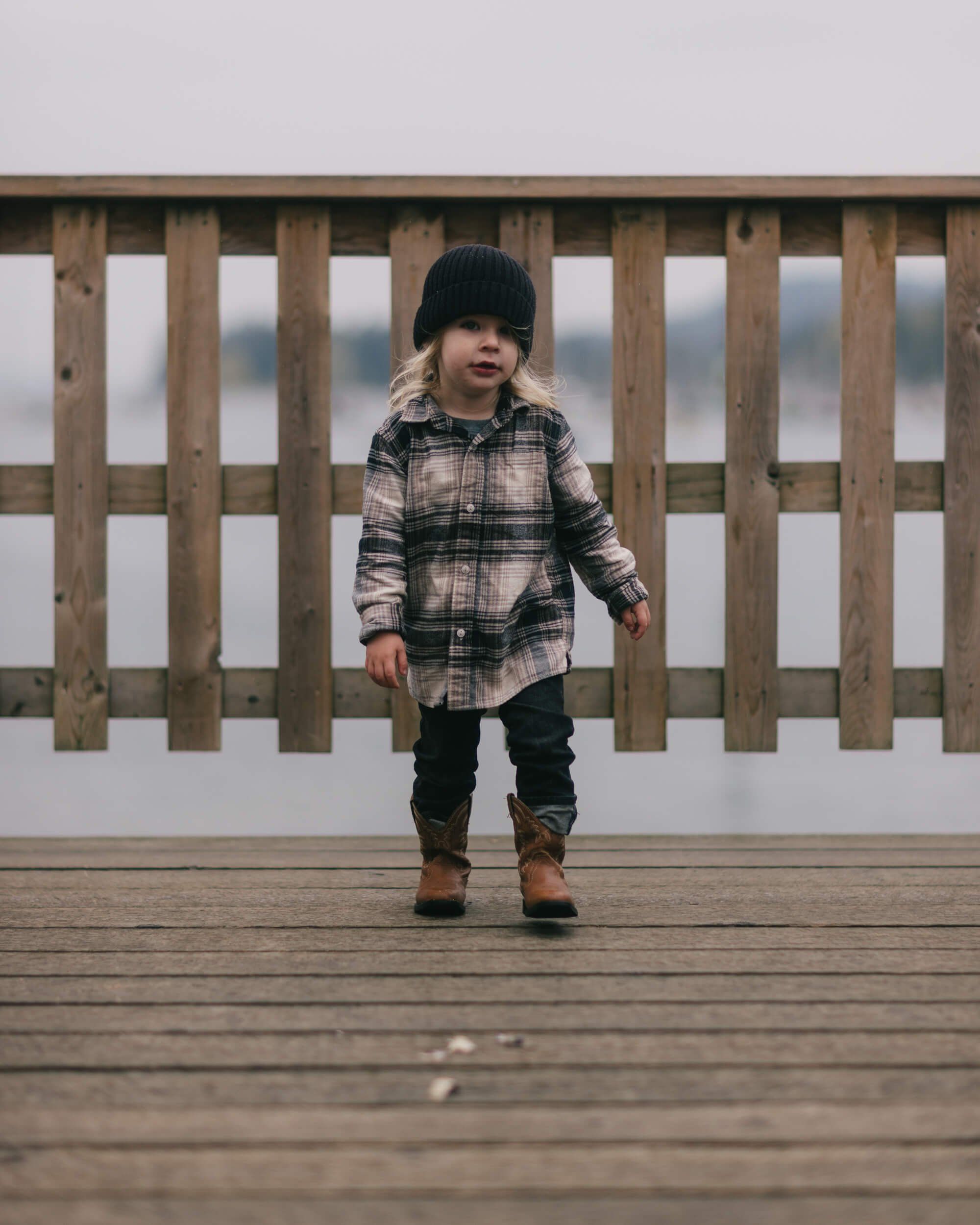 Toddler in plaid shirt and beanie stands on weathered wooden deck against overcast sky and railing