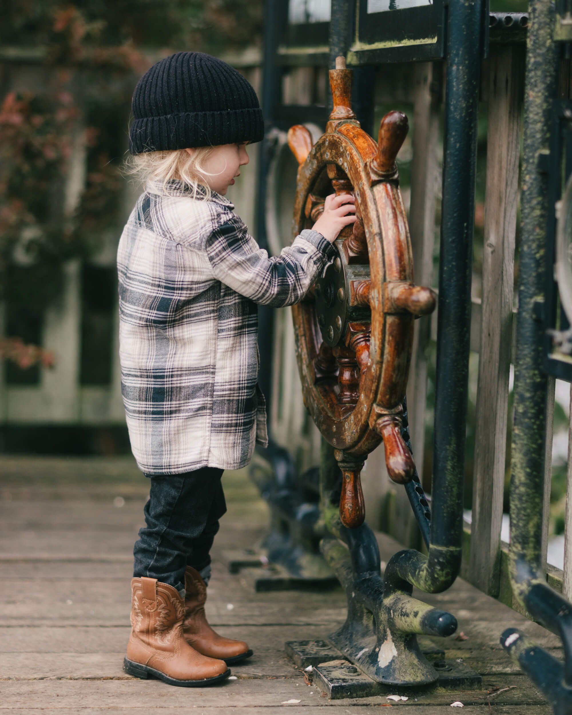 Young child in plaid shirt and black beanie examines wooden carved mask at outdoor market stall