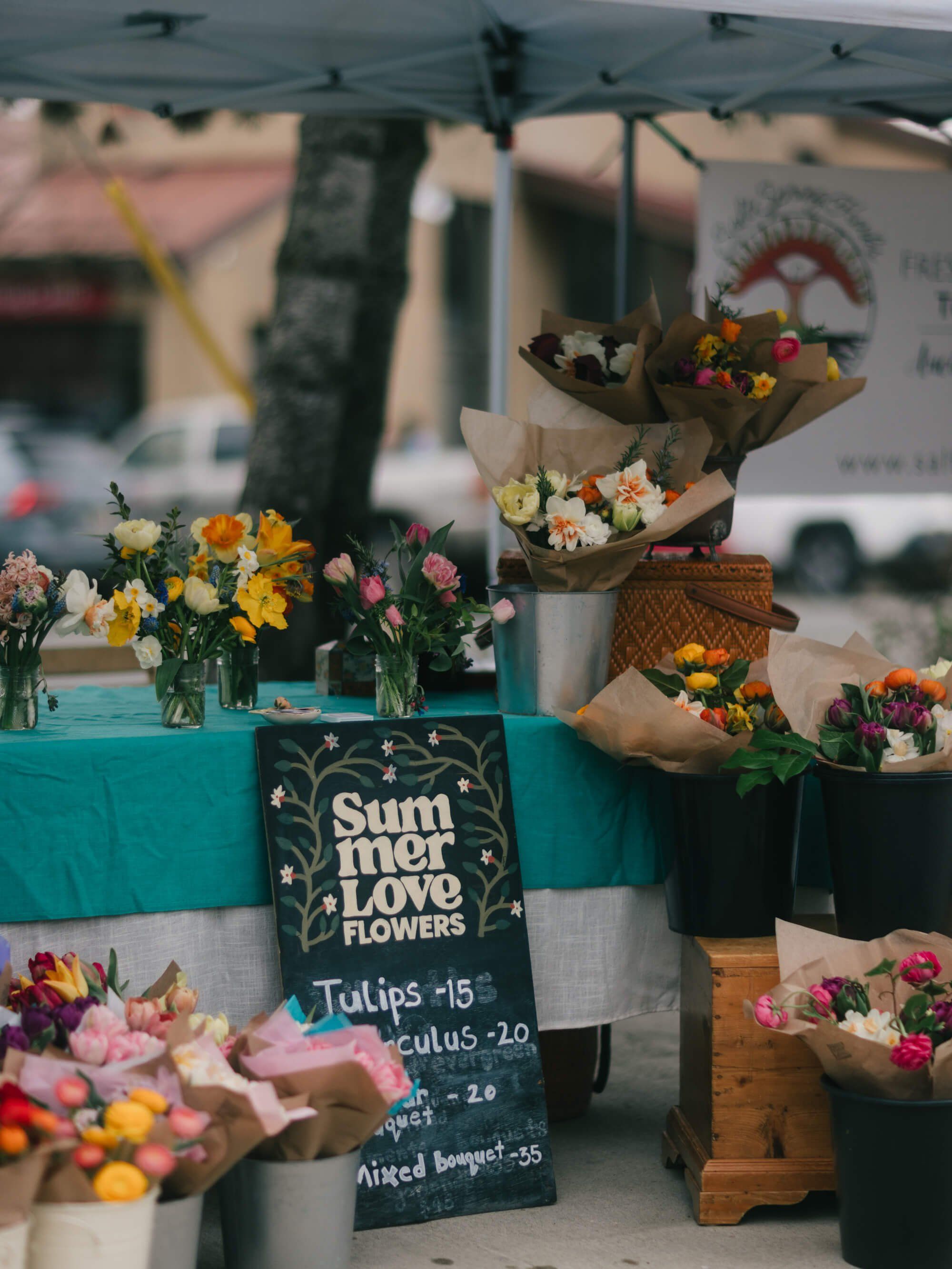 Flower vendor stall with turquoise fabric displaying colorful bouquets in vases and wrapped stems under market tent, warm afternoon light