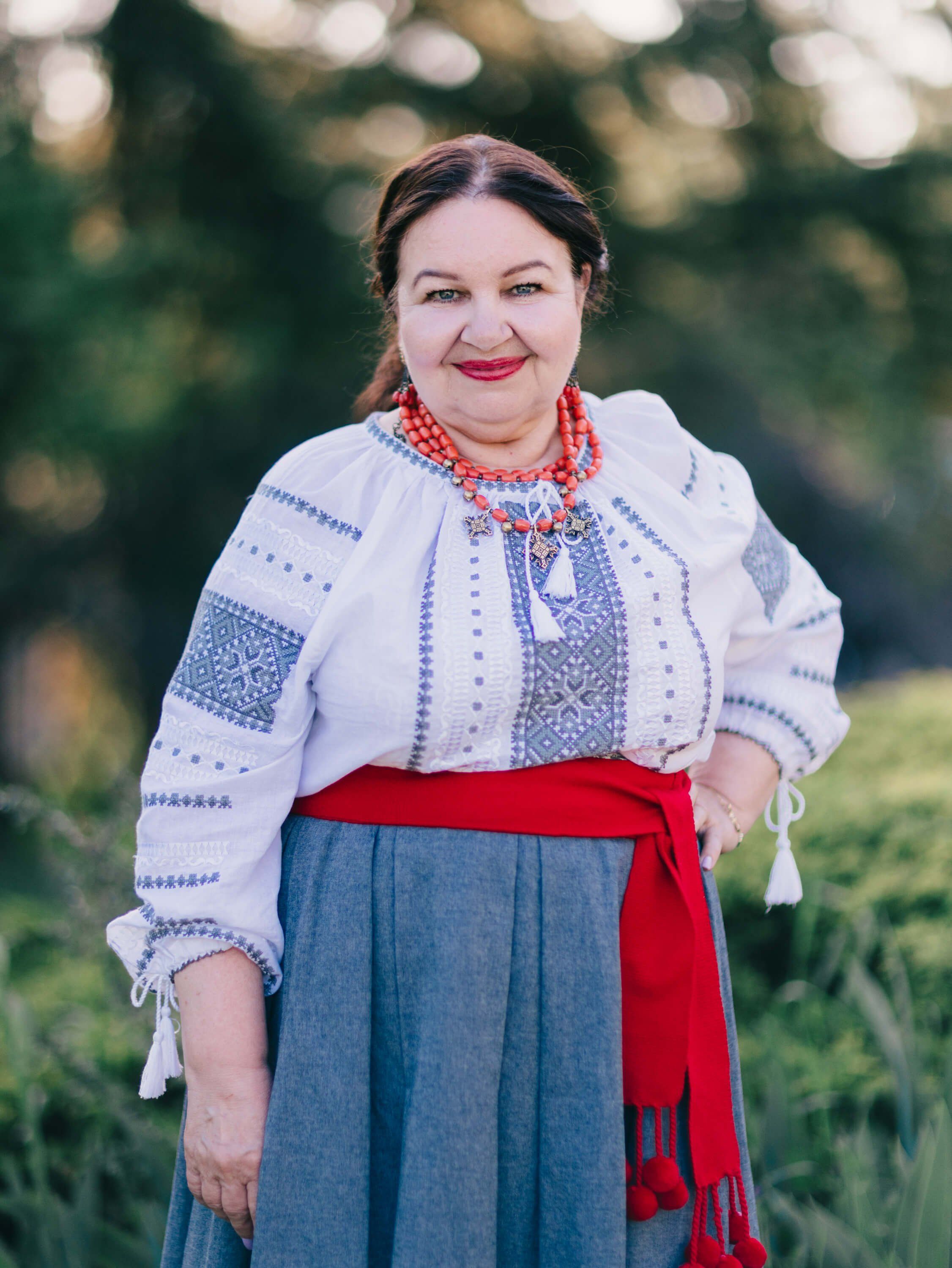 Woman in embroidered white blouse and blue skirt with red sash smiles warmly outdoors at golden hour