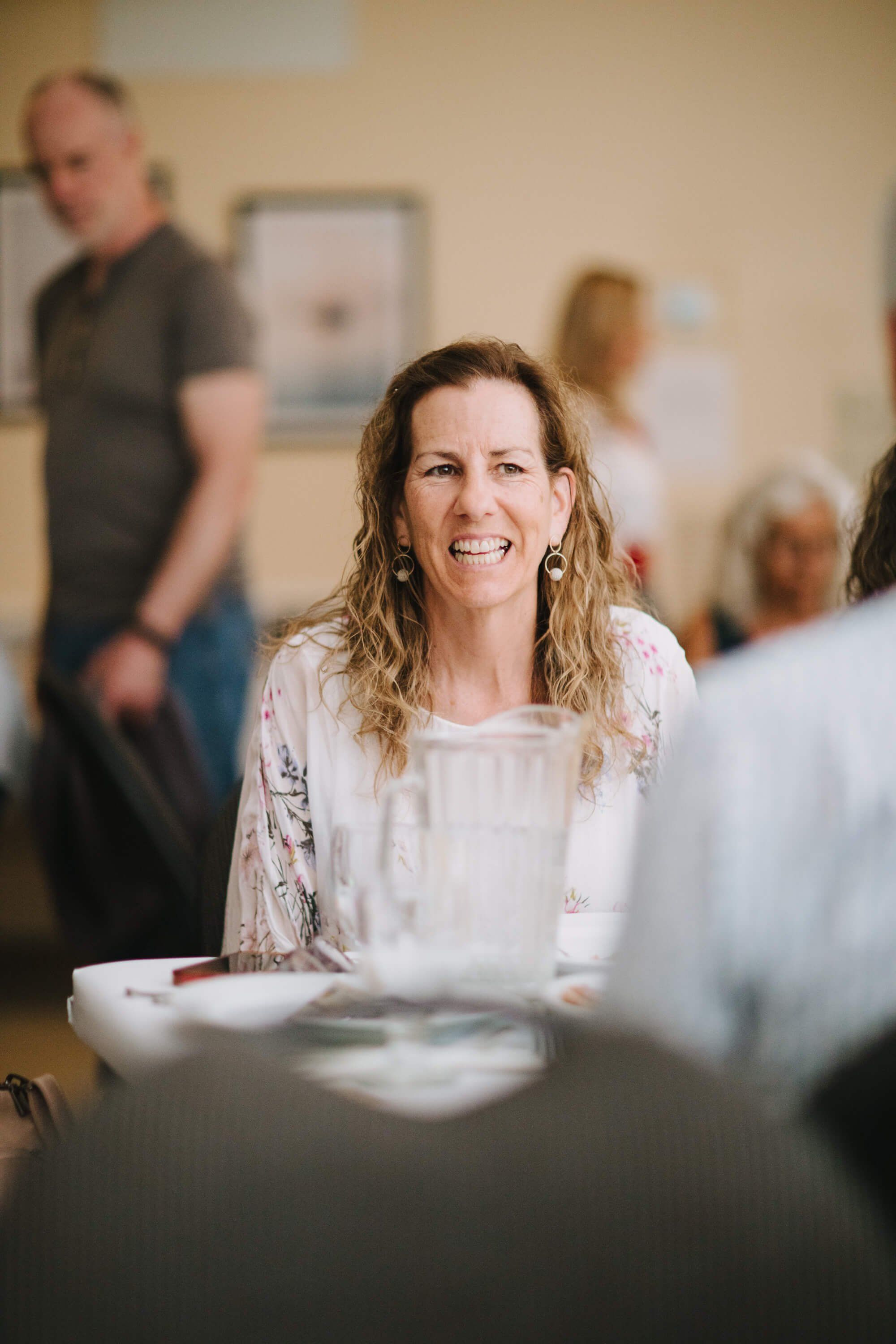 Woman with long wavy hair smiling warmly at a community dinner table, soft indoor lighting, blurred guests in background