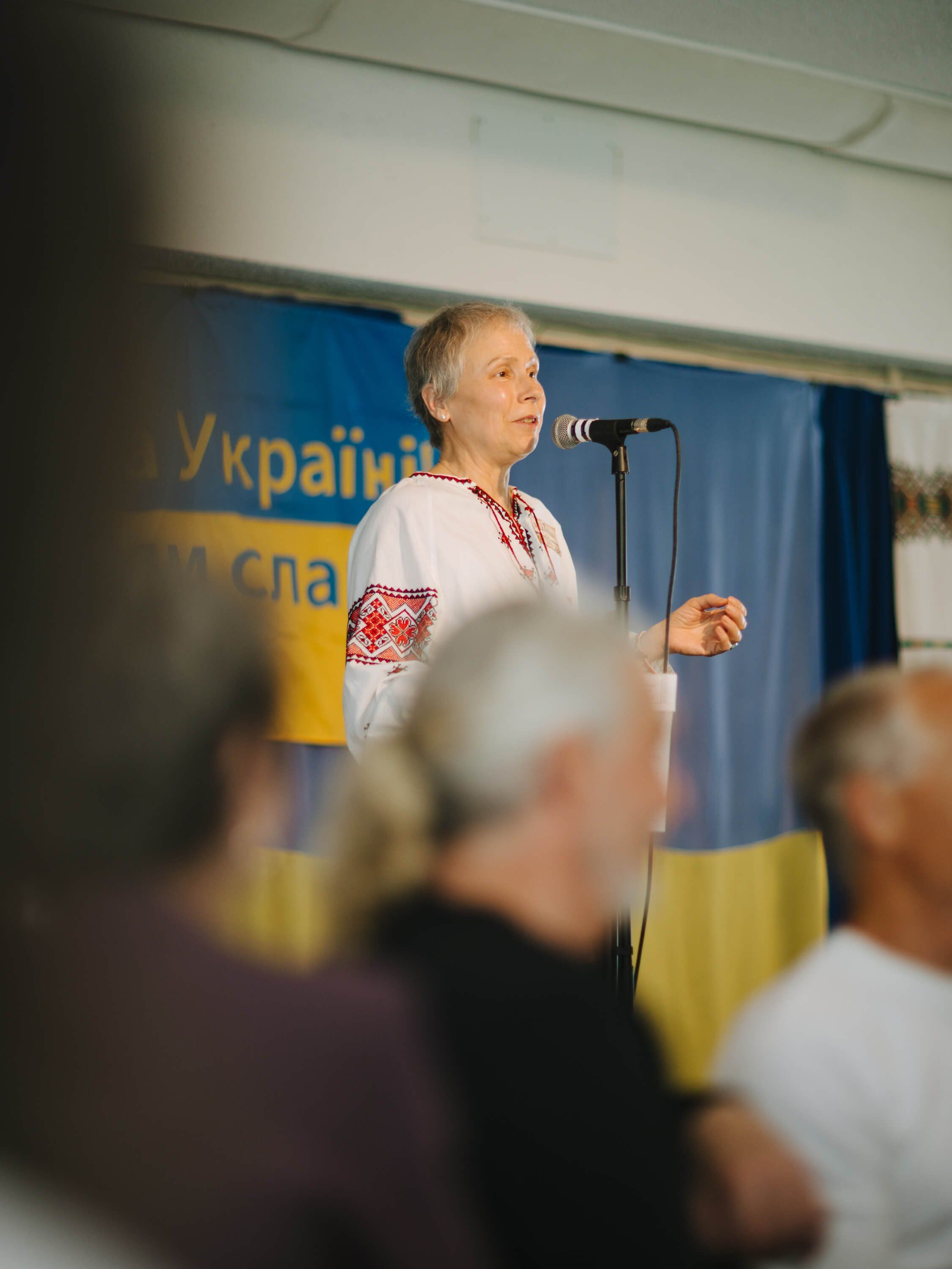 Man in embroidered Ukrainian shirt speaking at microphone before blue and yellow backdrop at fundraiser dinner