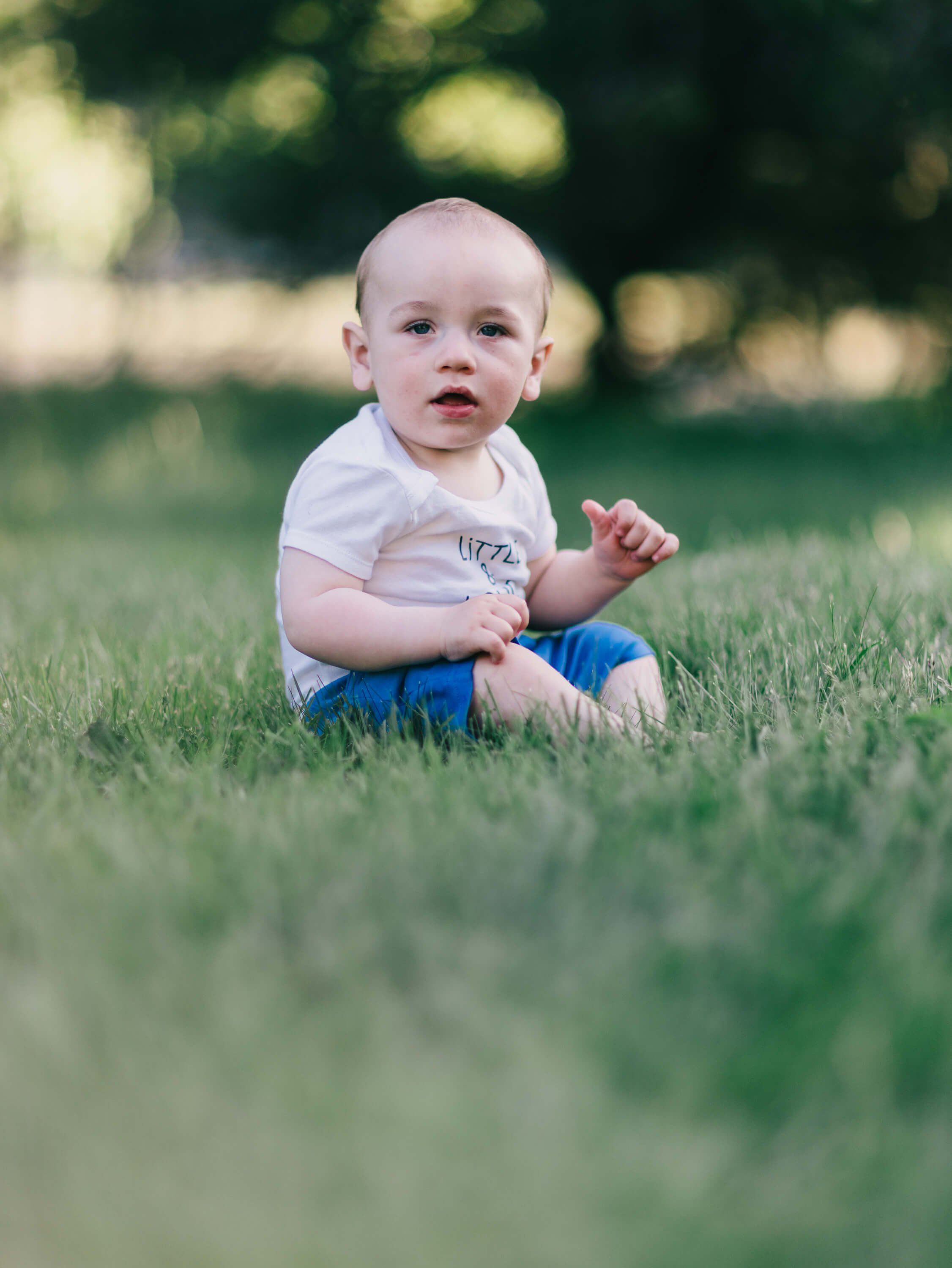 Blonde toddler in white shirt and blue pants sits on grass, gazing thoughtfully toward camera with soft afternoon light filtering through trees behind