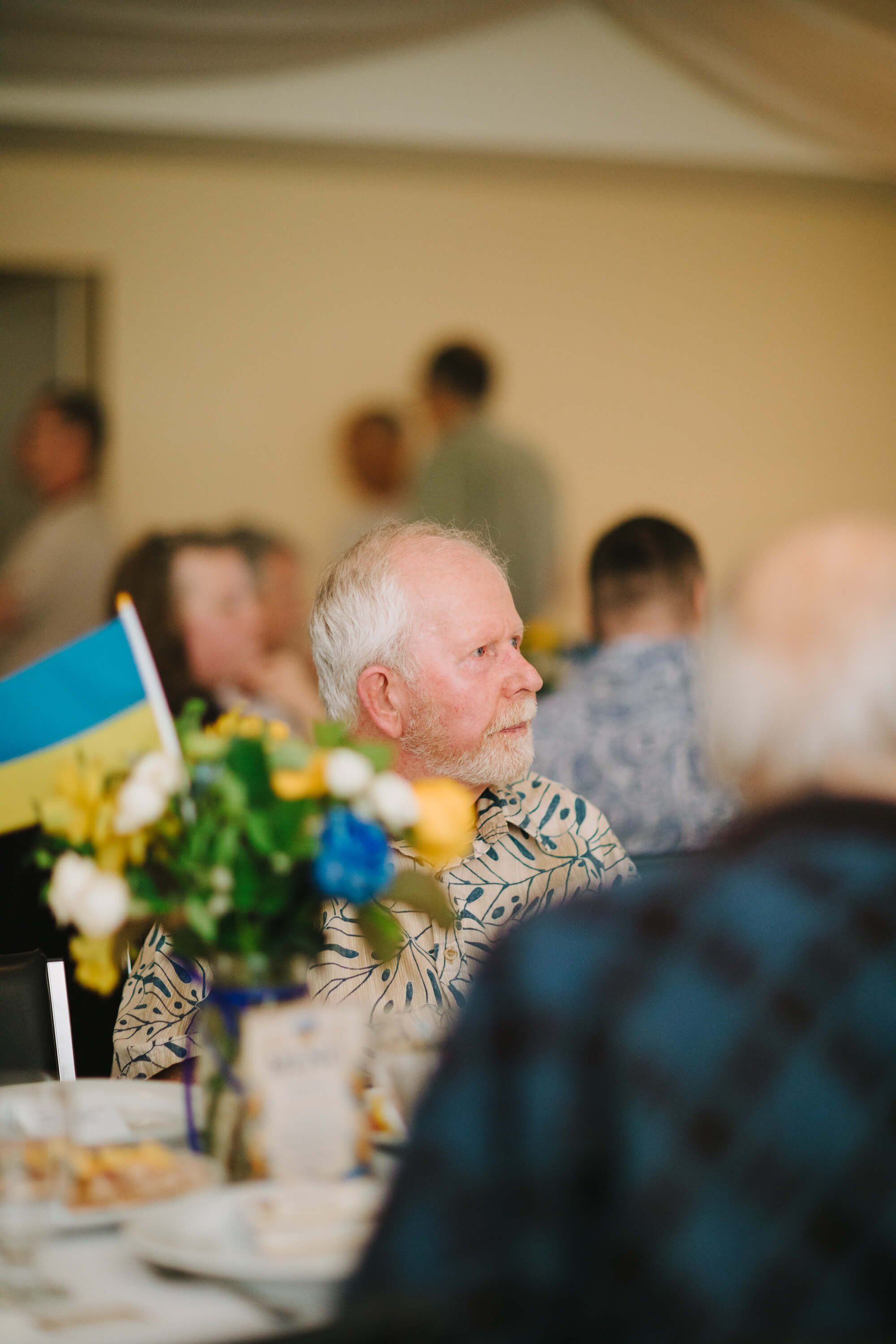 Elderly man in patterned shirt gazes thoughtfully beside Ukrainian flag and floral centerpiece at dinner event