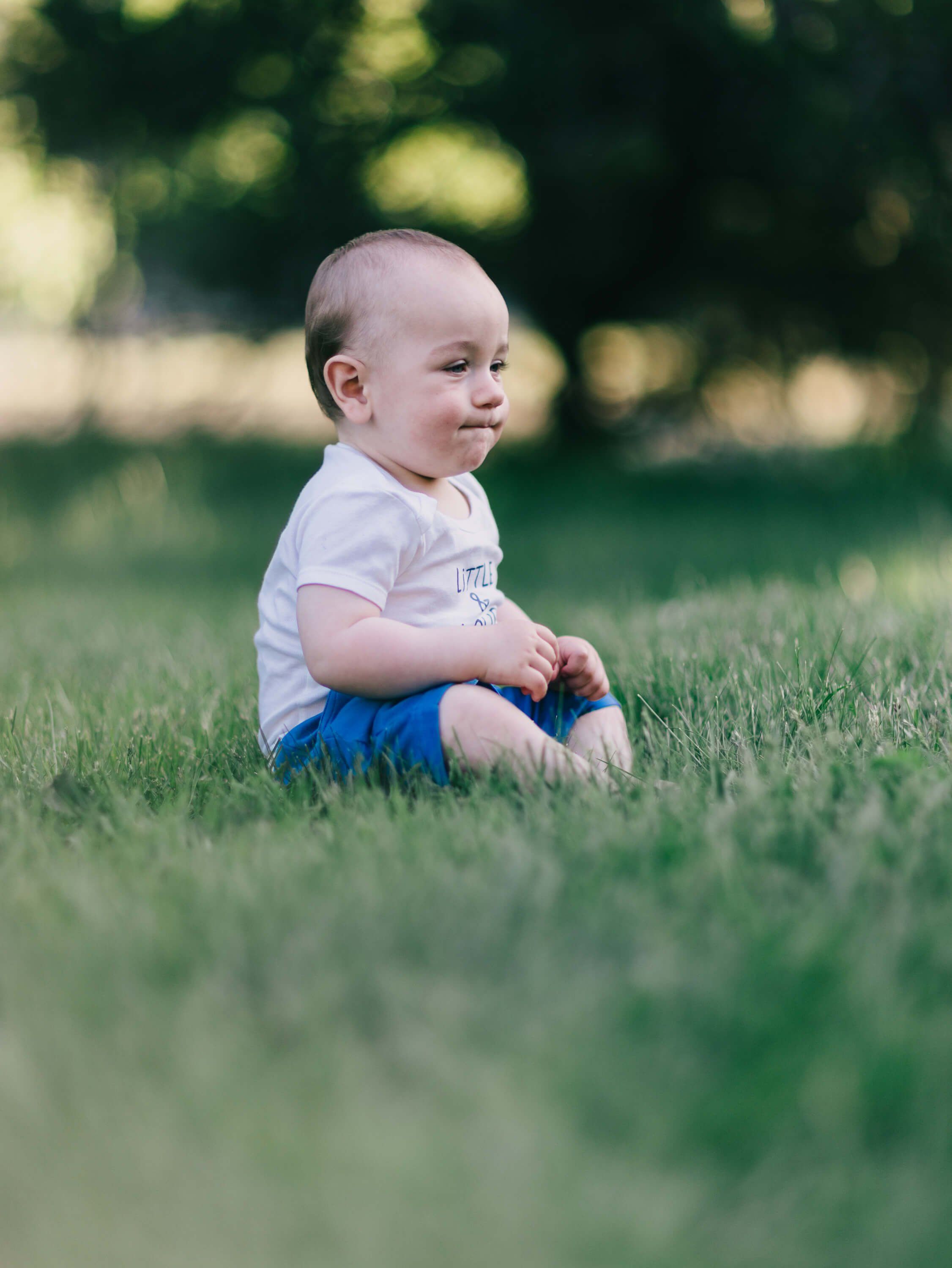 Blonde toddler in white shirt and blue shorts sits on grass, gazing thoughtfully to the side in golden afternoon light